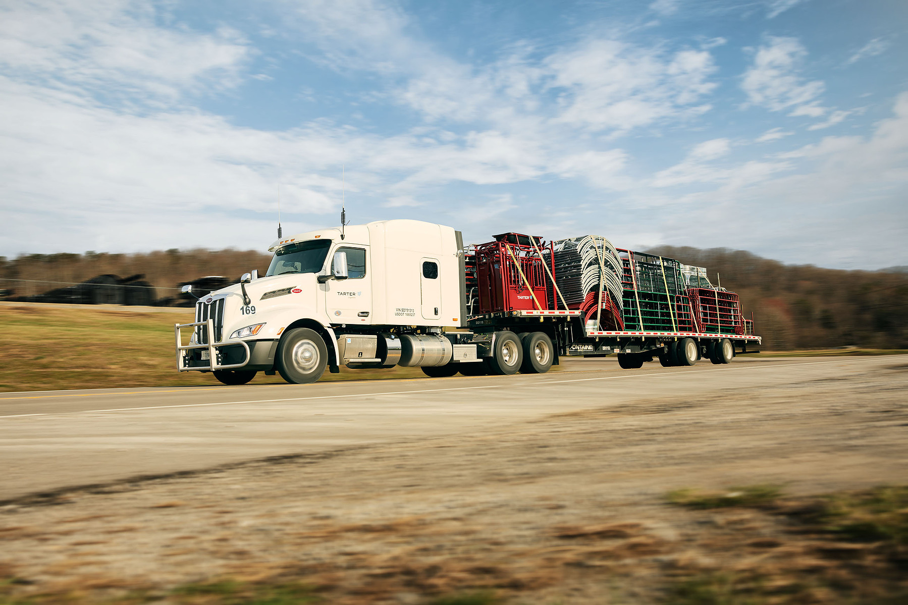 A white semi truck pulls a flatbed loaded with red and green farm equipment.