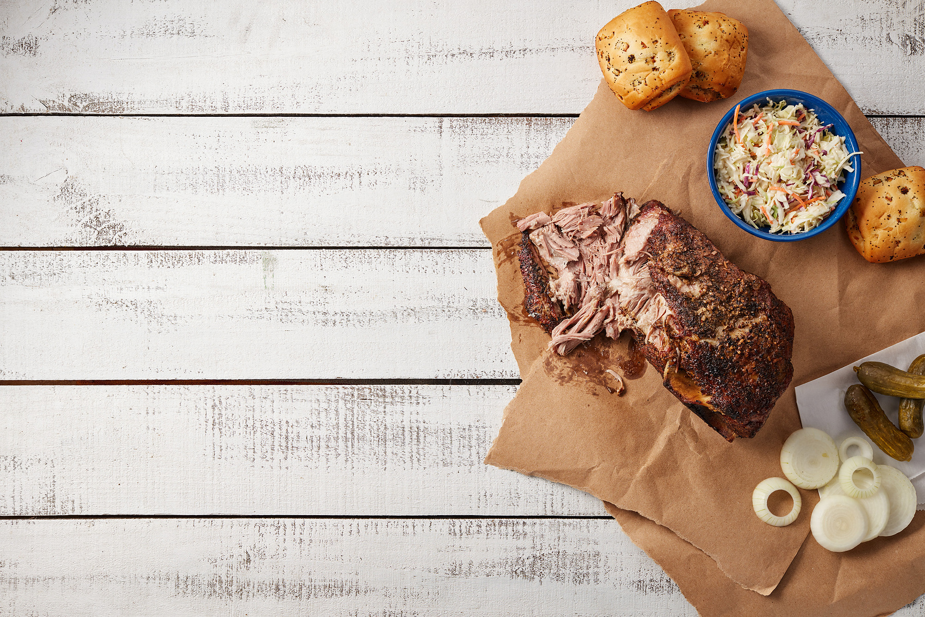 Pulled pork, a seasoned roast, bread rolls, coleslaw, pickles, and onion on brown serving paper.