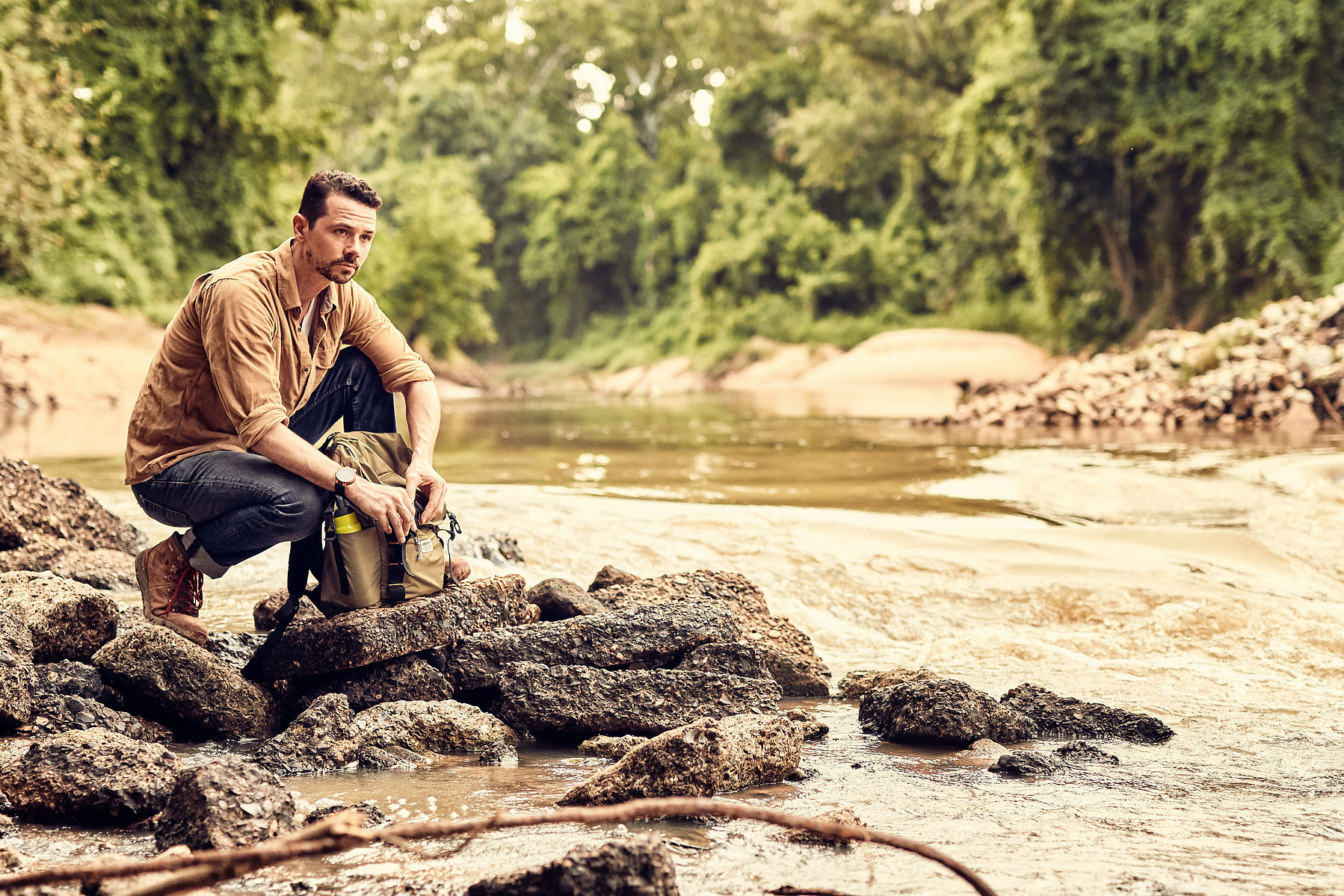 A man crouches on rocks beside a shallow creek in a wooded outdoor setting.