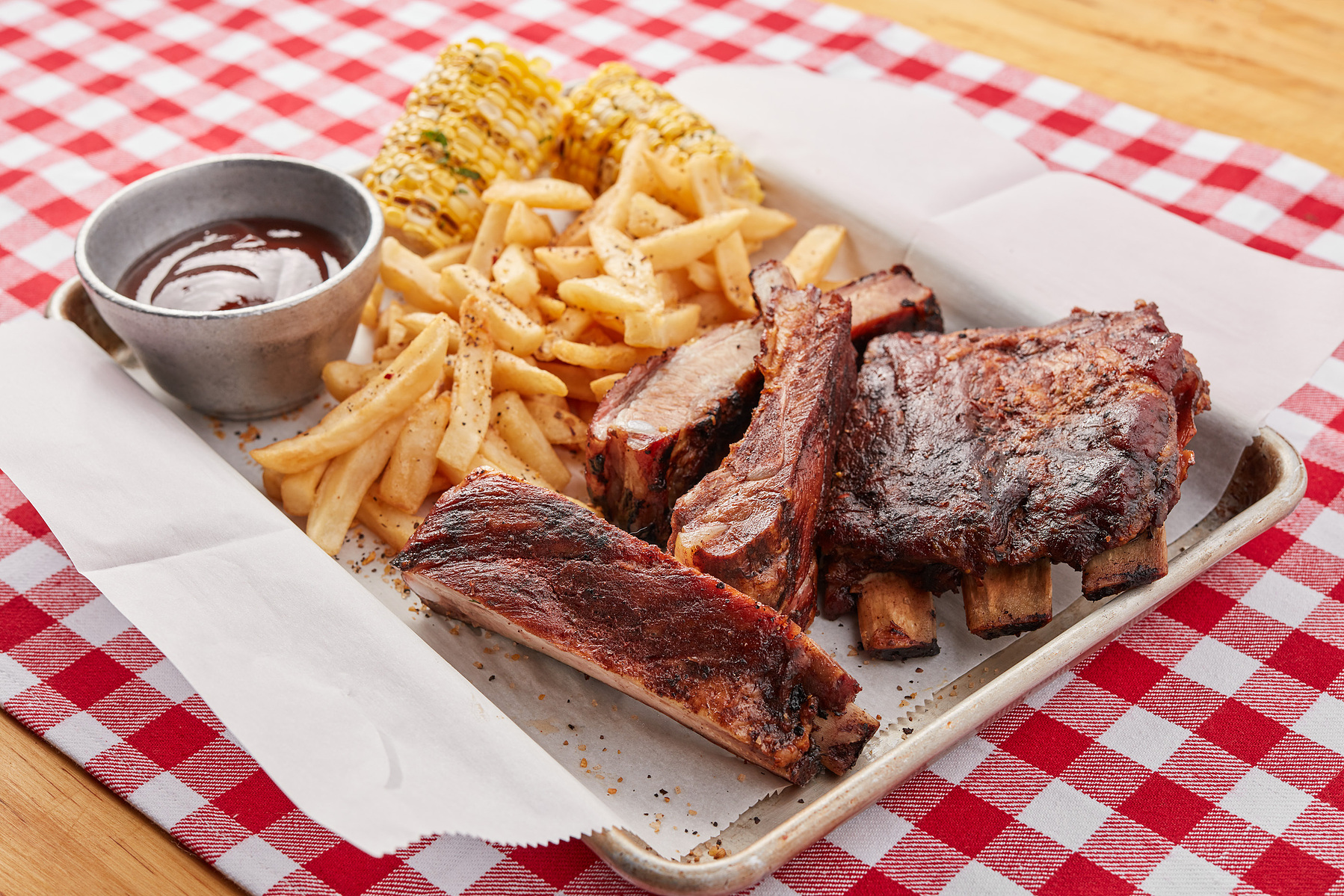 Barbecue ribs, sliced meat, fries, and sauce on a tray over red-and-white checked paper.