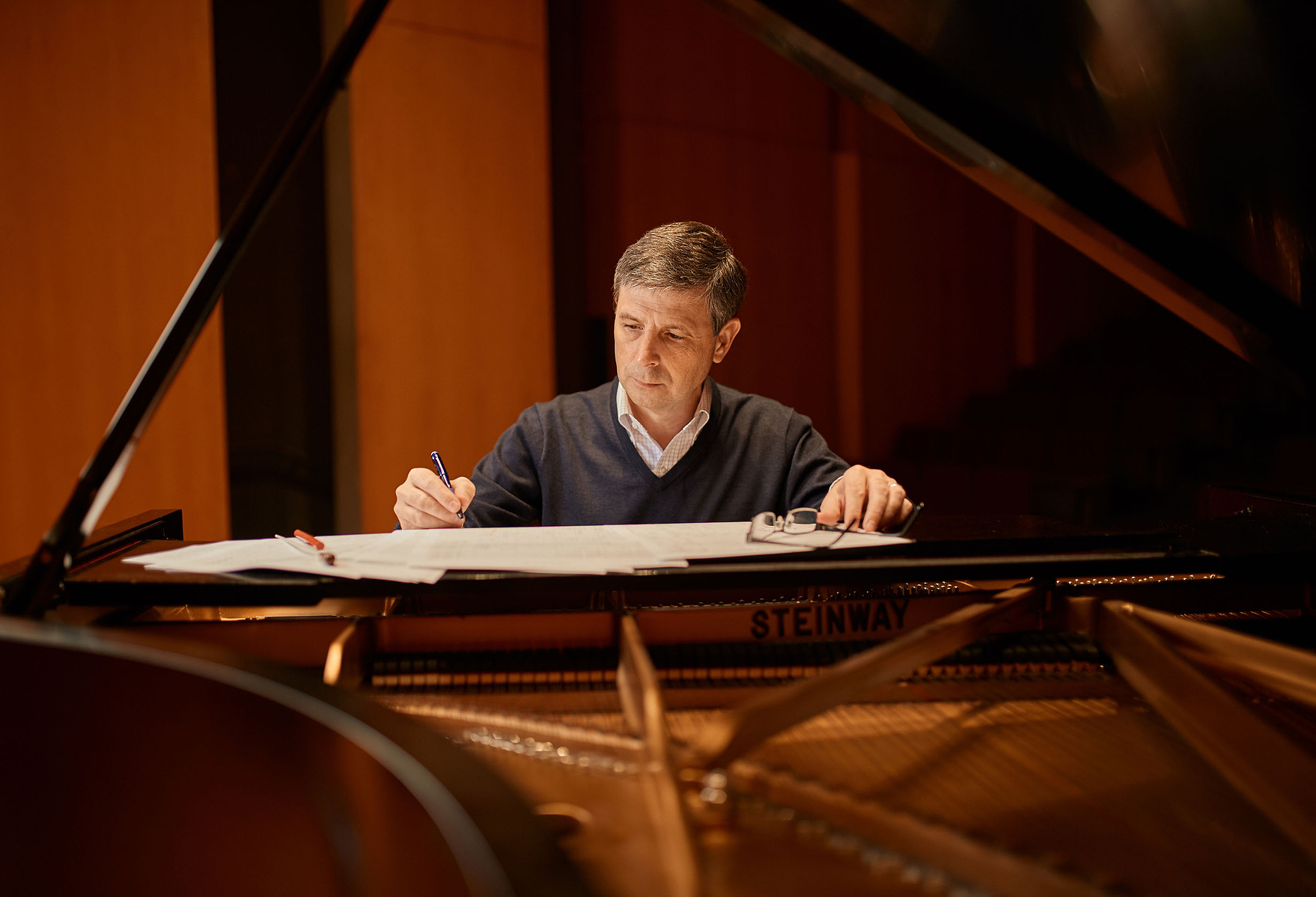 A man writes on sheet music beside the open lid of a grand piano in warm indoor light.