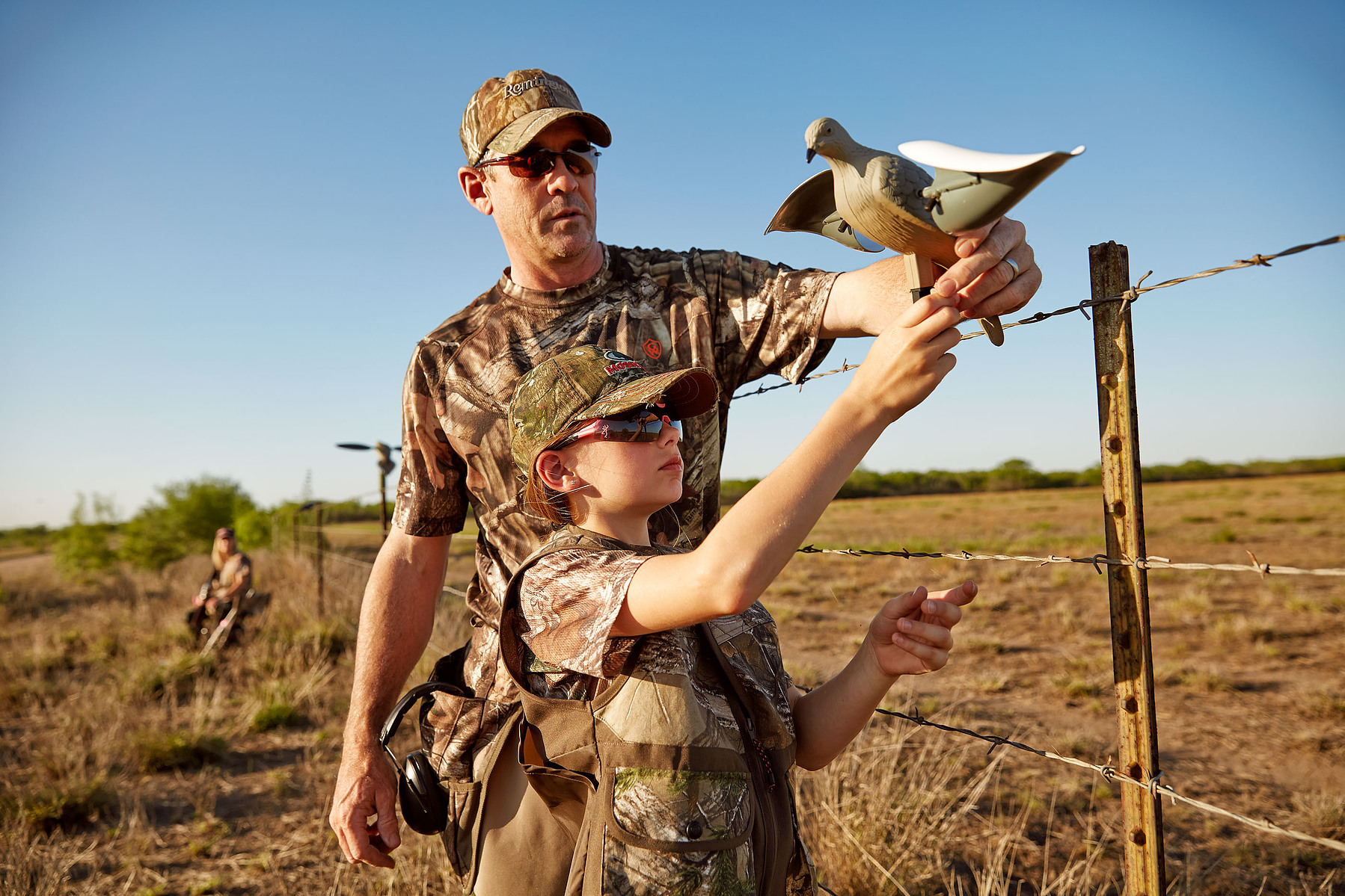 An adult and child wearing camouflage position a bird decoy beside a wire fence.