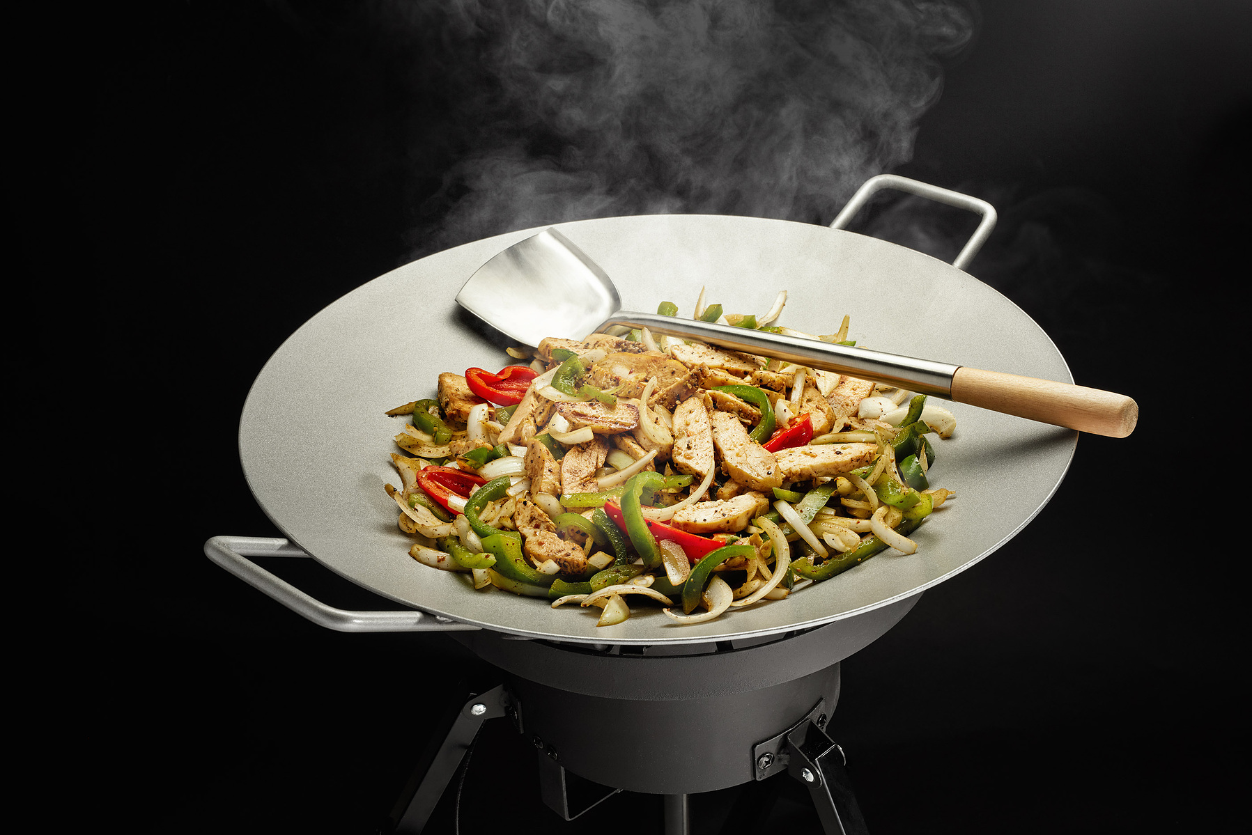 Strips of meat and colorful vegetables steam in a wok with a wooden-handled spatula.