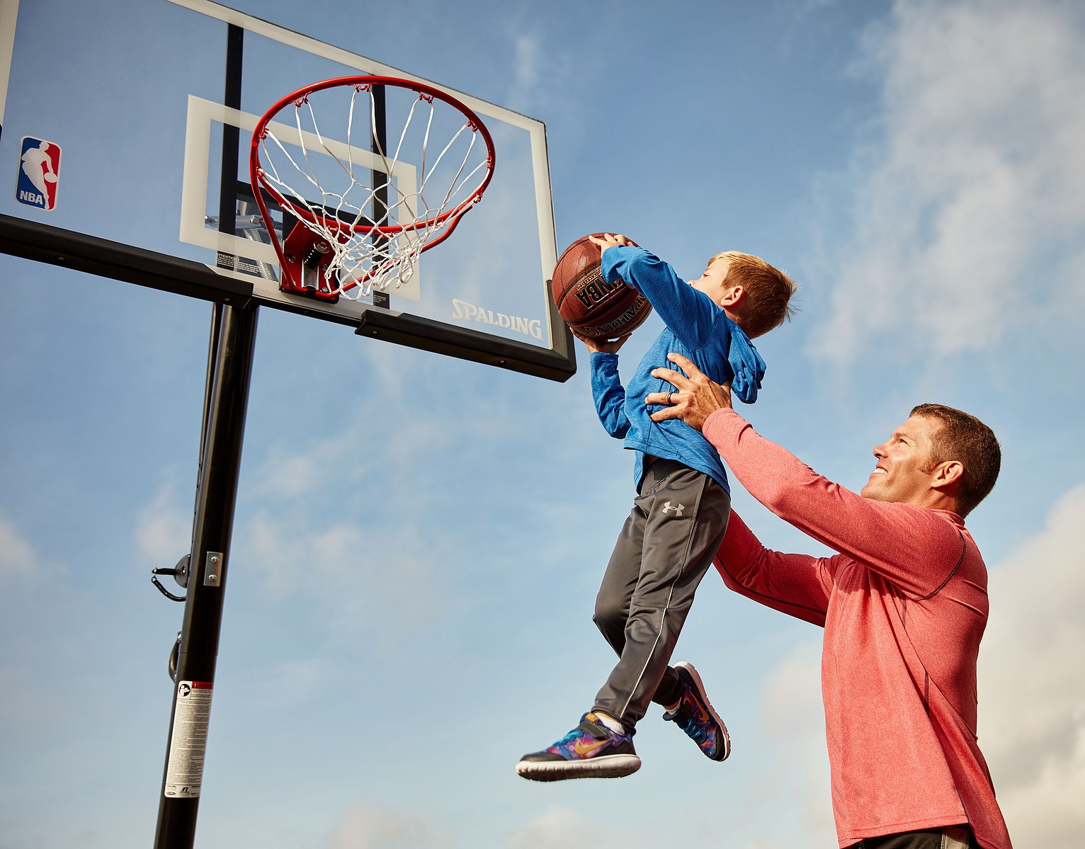 An adult lifts a child holding a basketball toward an outdoor basketball hoop.