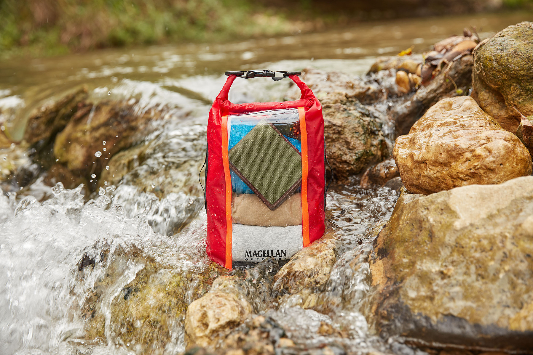 A red Magellan dry bag rests on rocks beside flowing creek water.