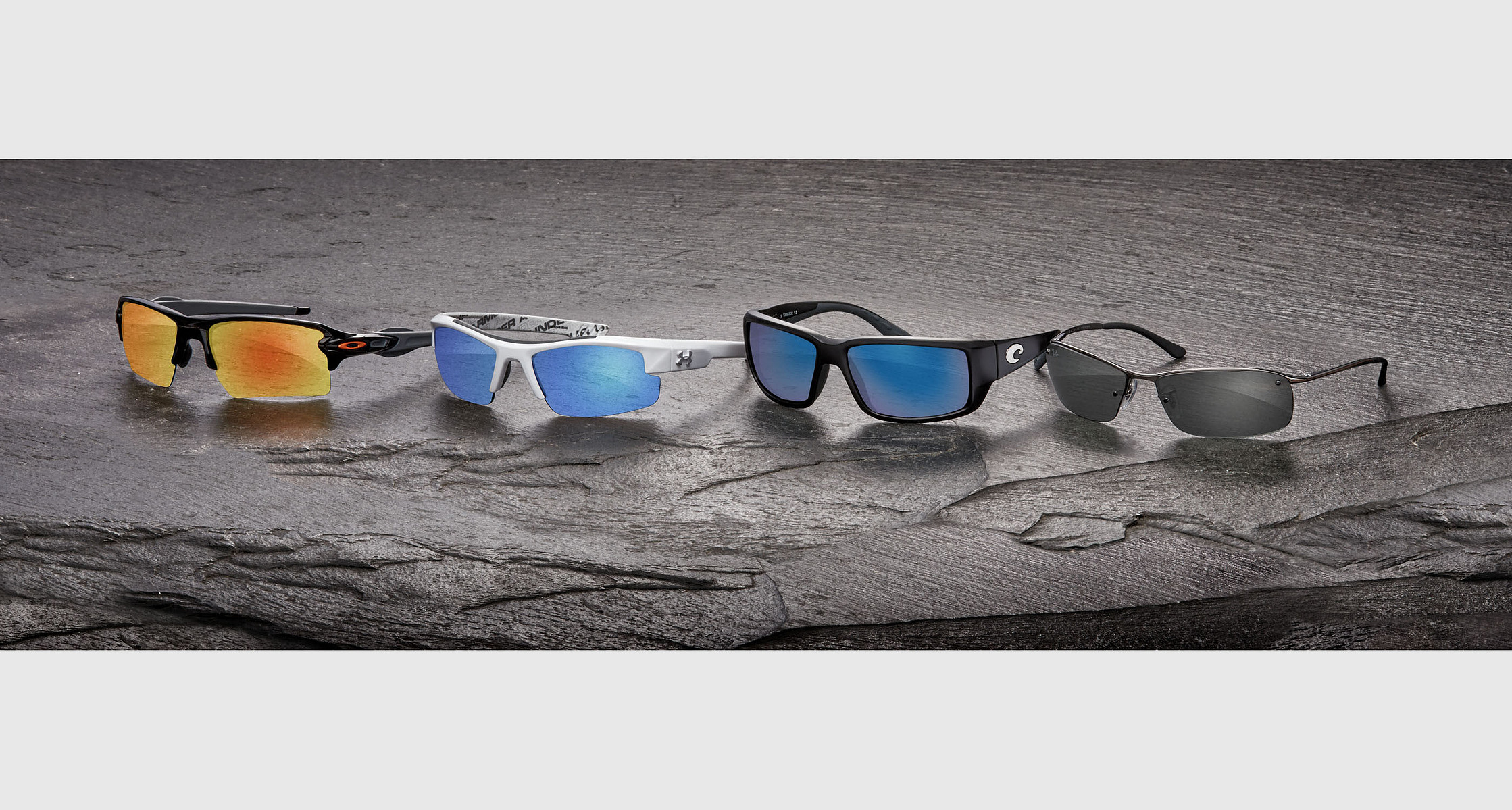 Four pairs of sport sunglasses with colored lenses arranged on a textured gray surface.