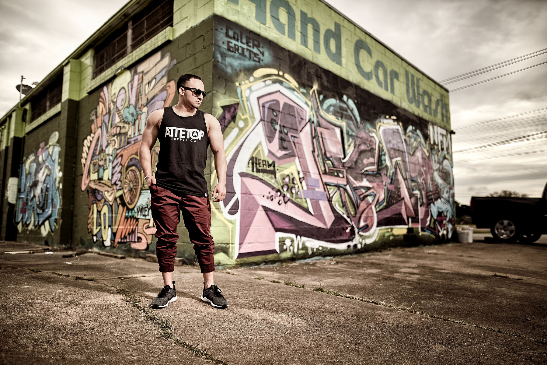 A man in sunglasses and athletic streetwear stands beside a graffiti-covered industrial building.