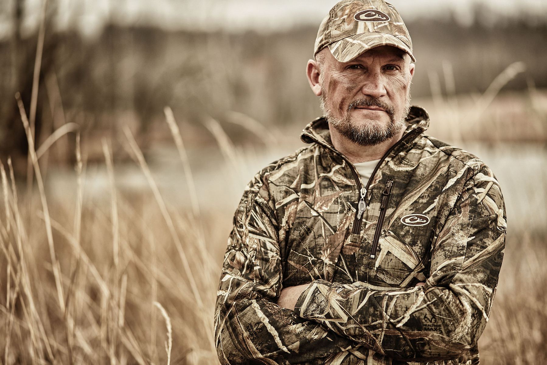 A man in a camouflage jacket and cap stands with his arms folded in a reed-filled landscape.