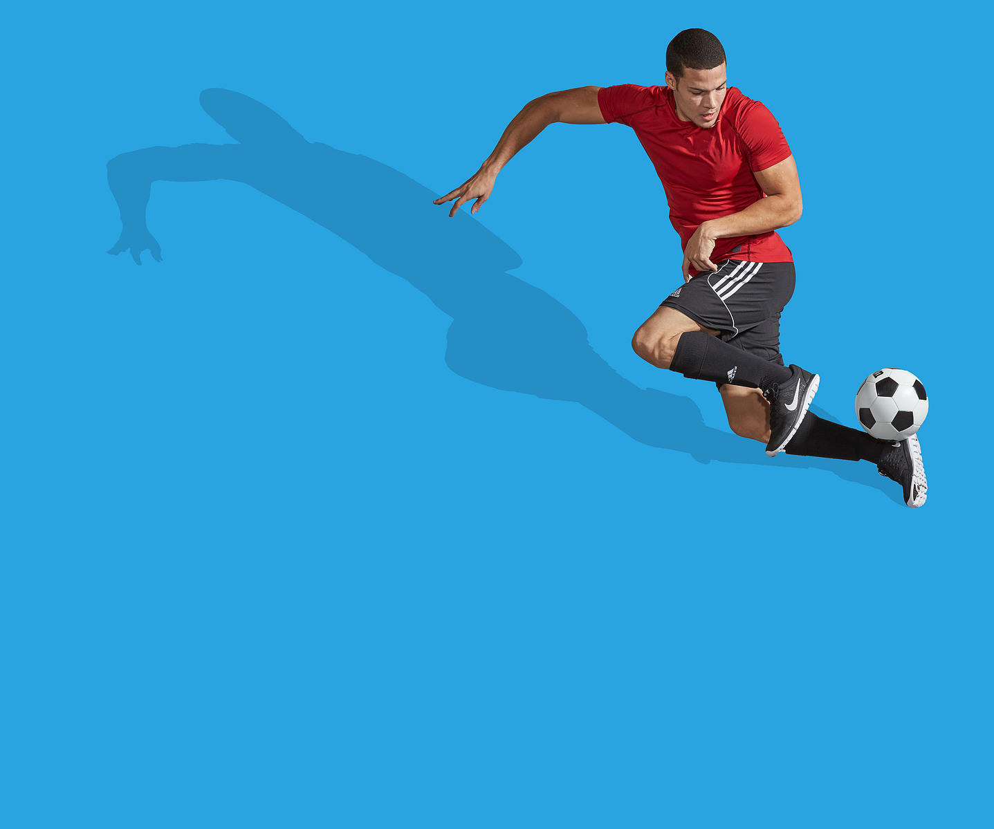 A soccer player in a red shirt jumps beside a ball against a bright blue studio background.