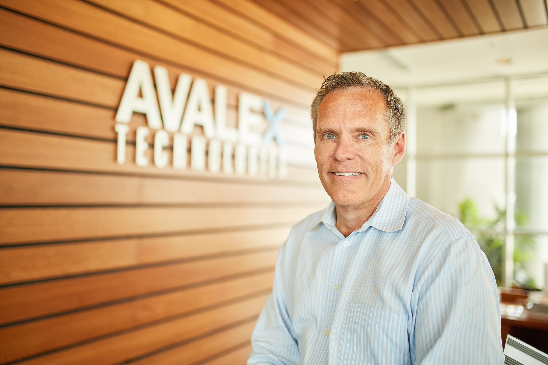 A man in a pale striped shirt stands beside an Avalex Technologies sign on a wooden wall.