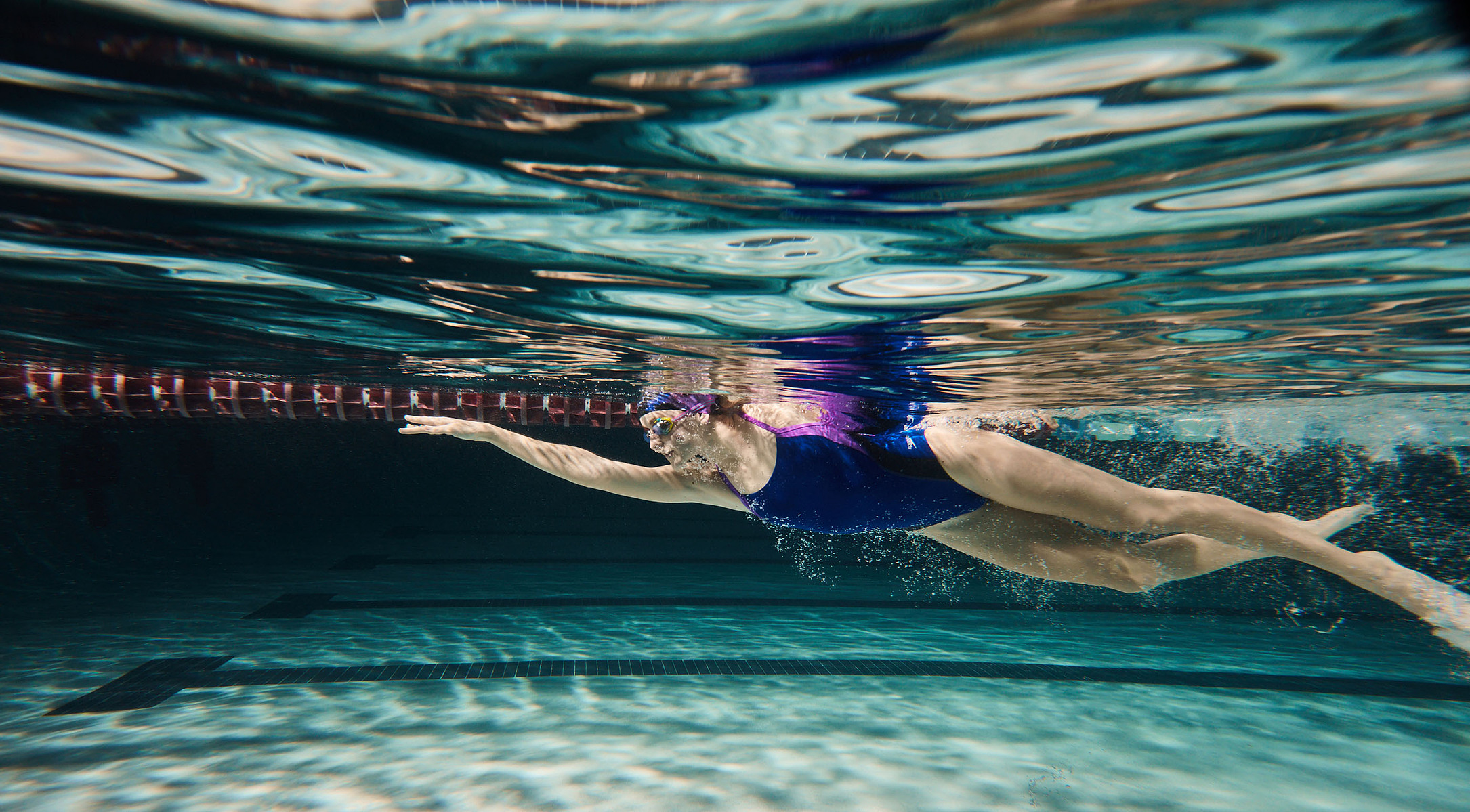 An underwater view of a swimmer moving through a pool beneath rippling reflections.