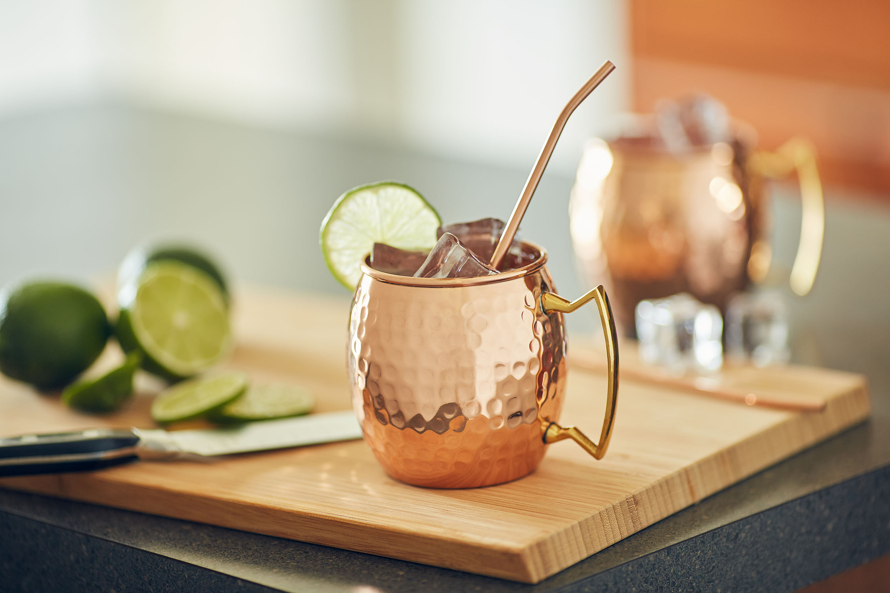 A copper drink mug with ice, lime, and a metal straw on a wooden board beside whole limes.