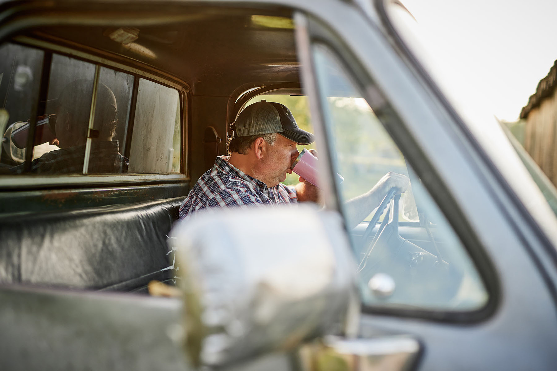 A farmer sits in the cab of an older pickup truck, viewed through the side window.