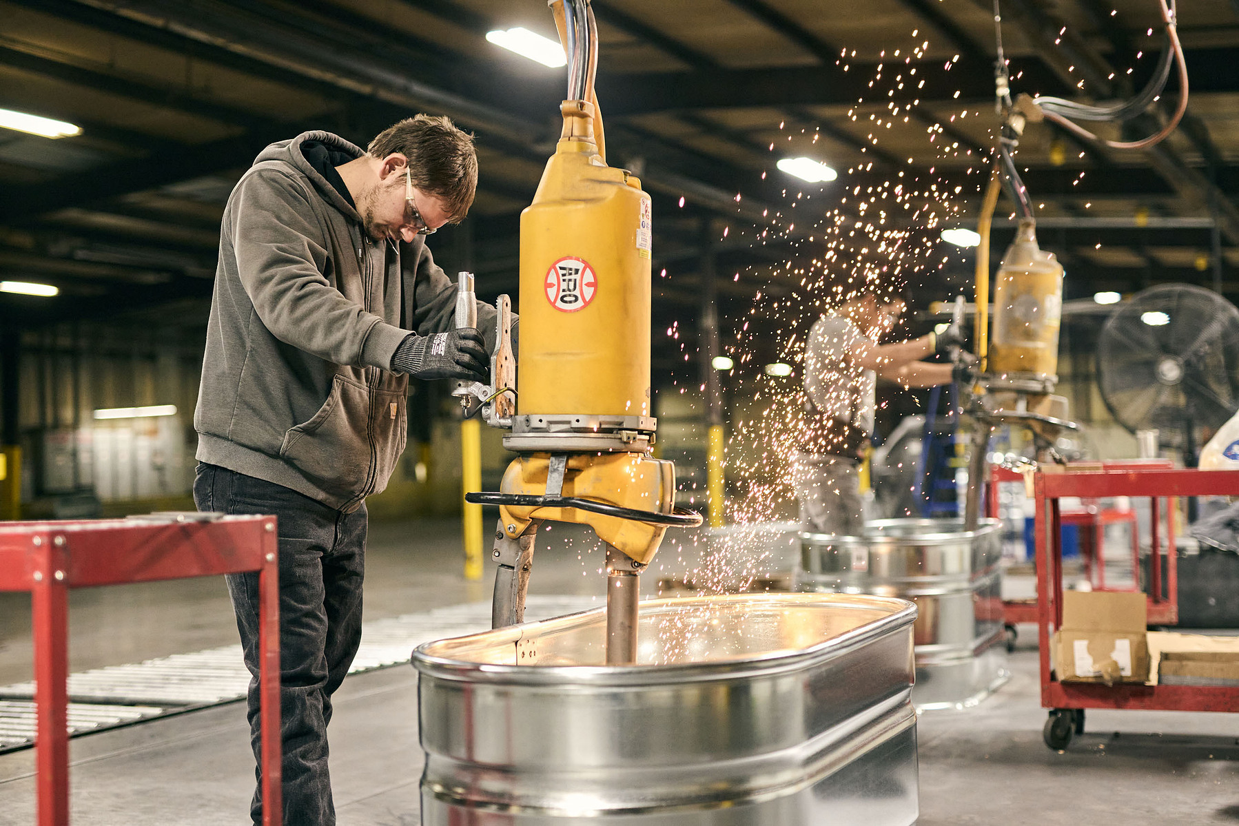 A worker operates a yellow machine beside a galvanized stock tank as sparks scatter.