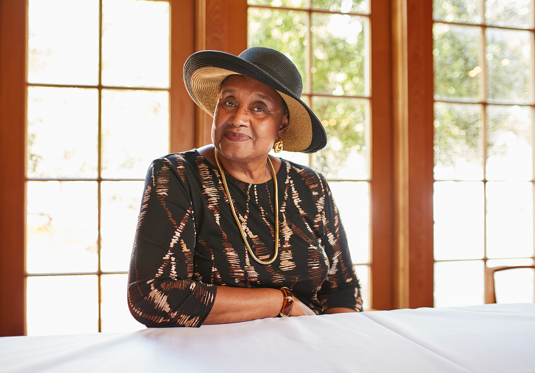 A woman wearing a dark hat and layered necklaces sits at a table beside large windows.