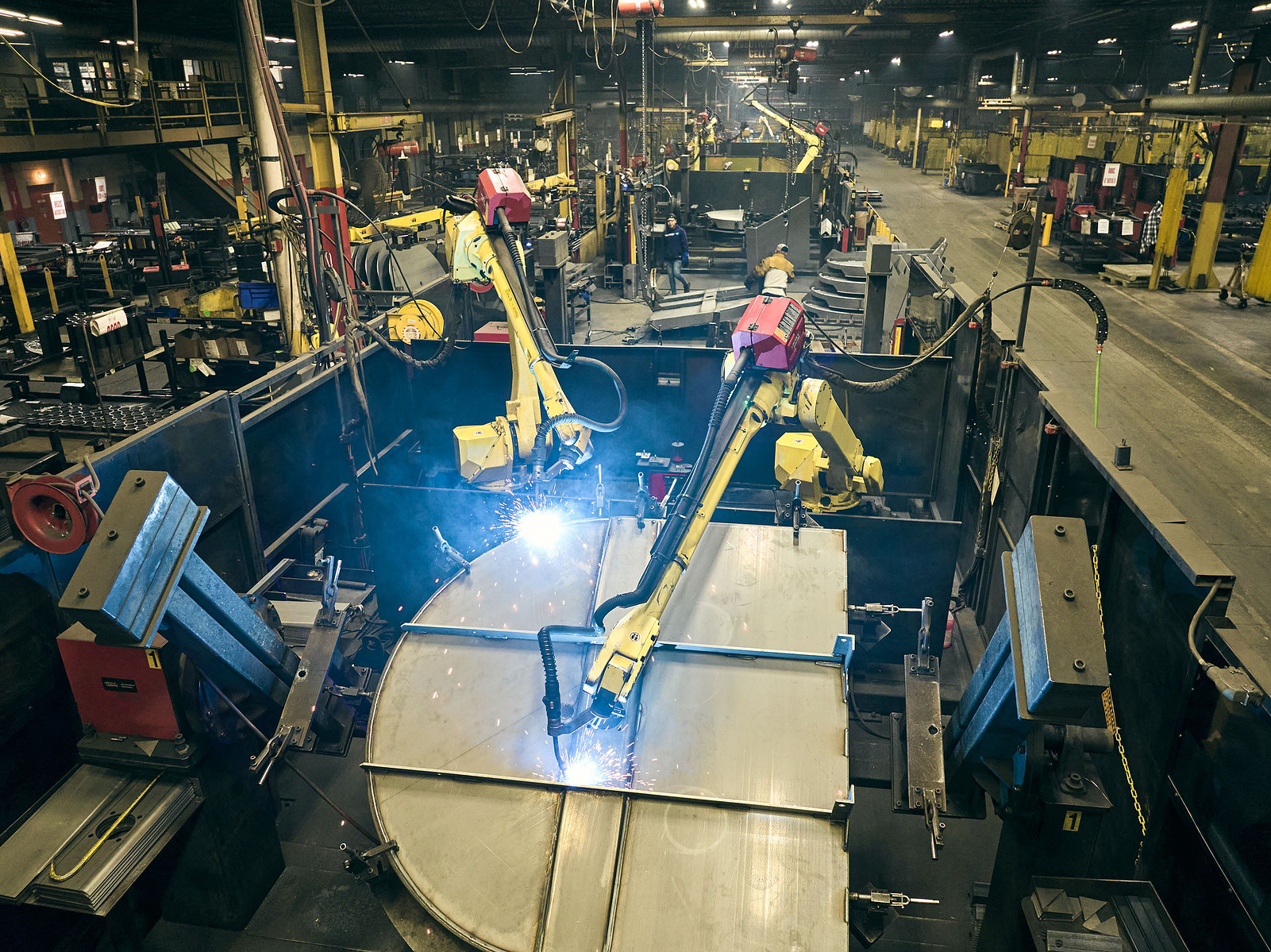 An elevated view shows yellow robotic welding arms and workers across a busy factory floor.