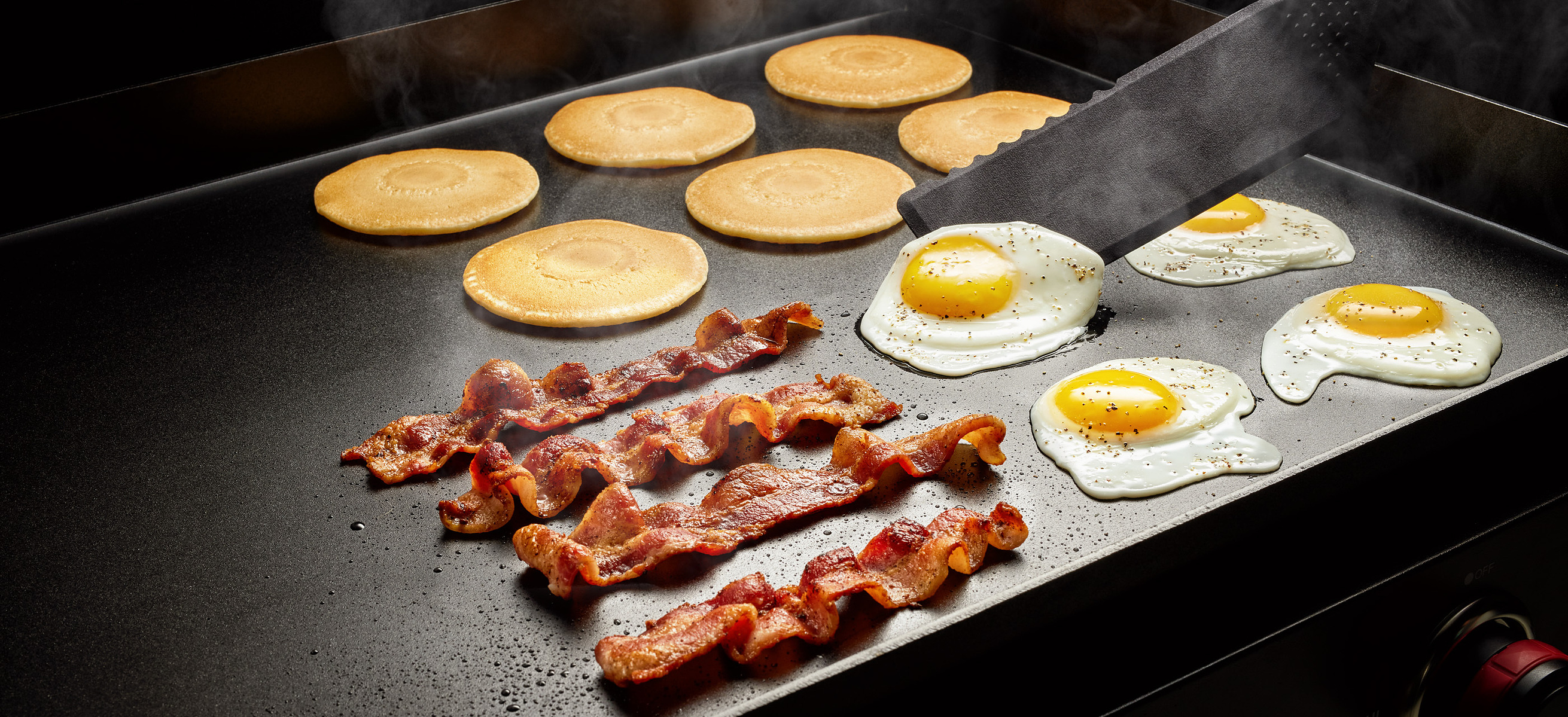 Pancakes, bacon strips, and fried eggs cook on a flat black griddle.