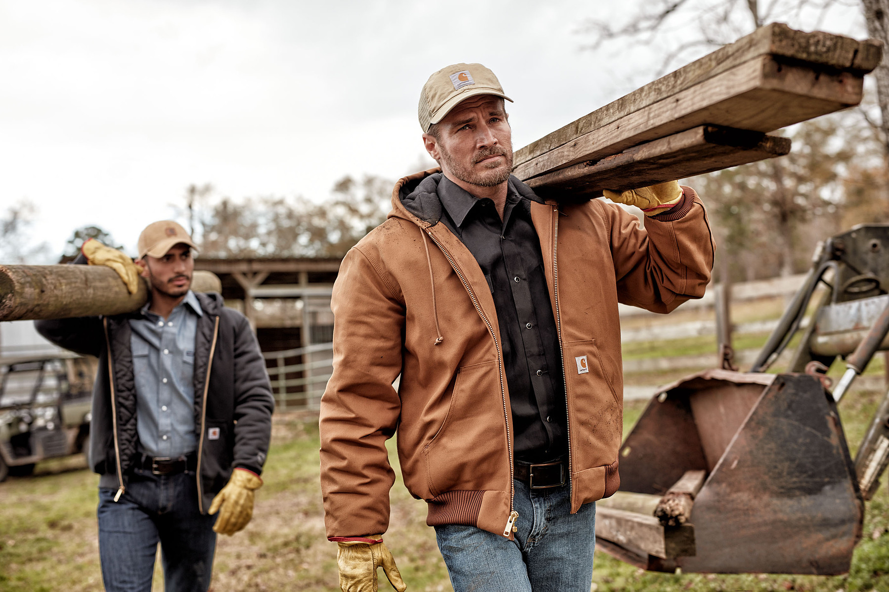 Two people in work jackets carry timber through an outdoor work area.