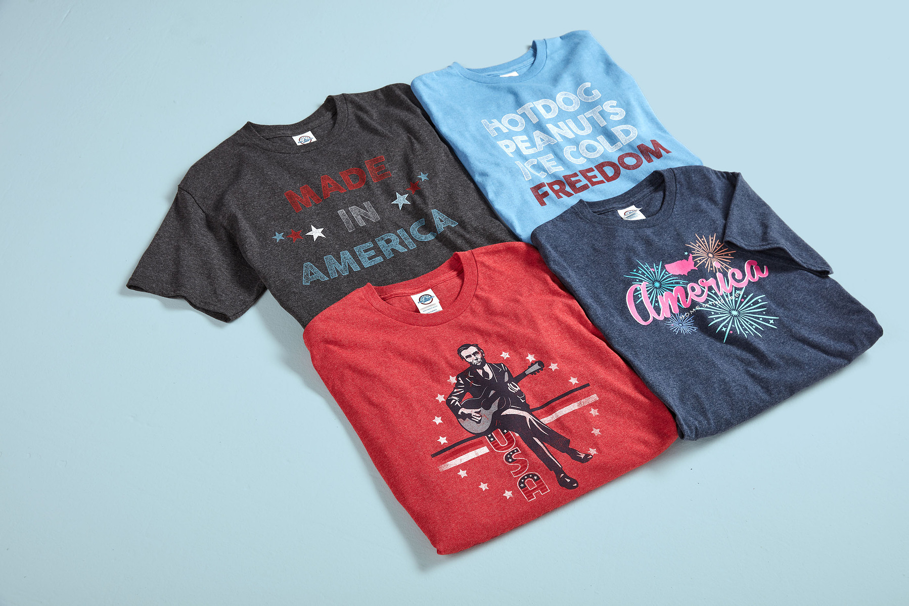 Four graphic T-shirts in black, blue, red, and navy arranged on a pale blue background.