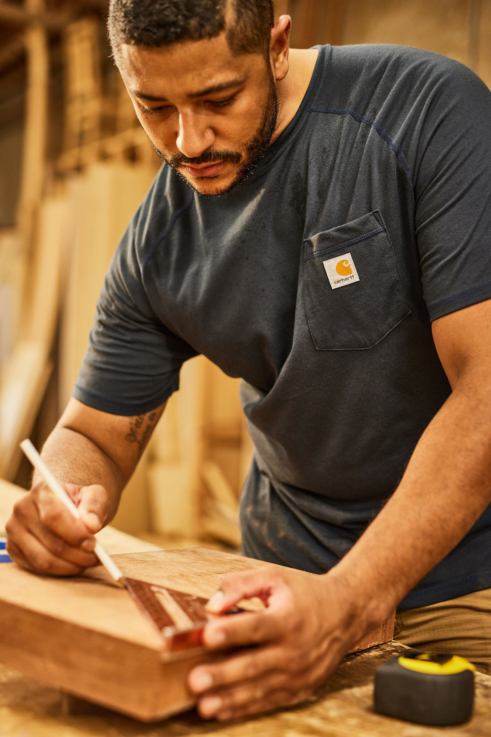 A man wearing a dark work shirt marks a wooden board with a pencil in a workshop.