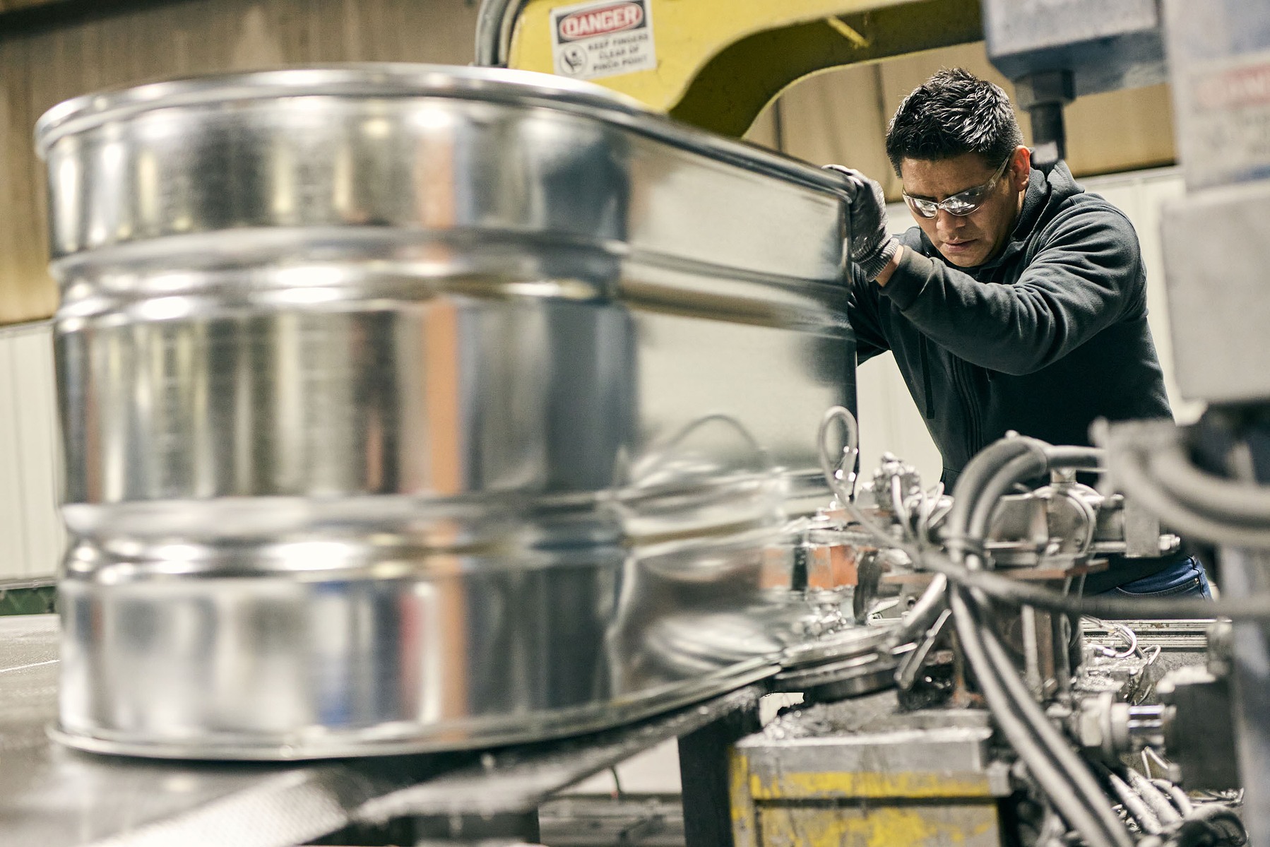 A worker wearing safety glasses handles a polished galvanized tank beside industrial machinery.