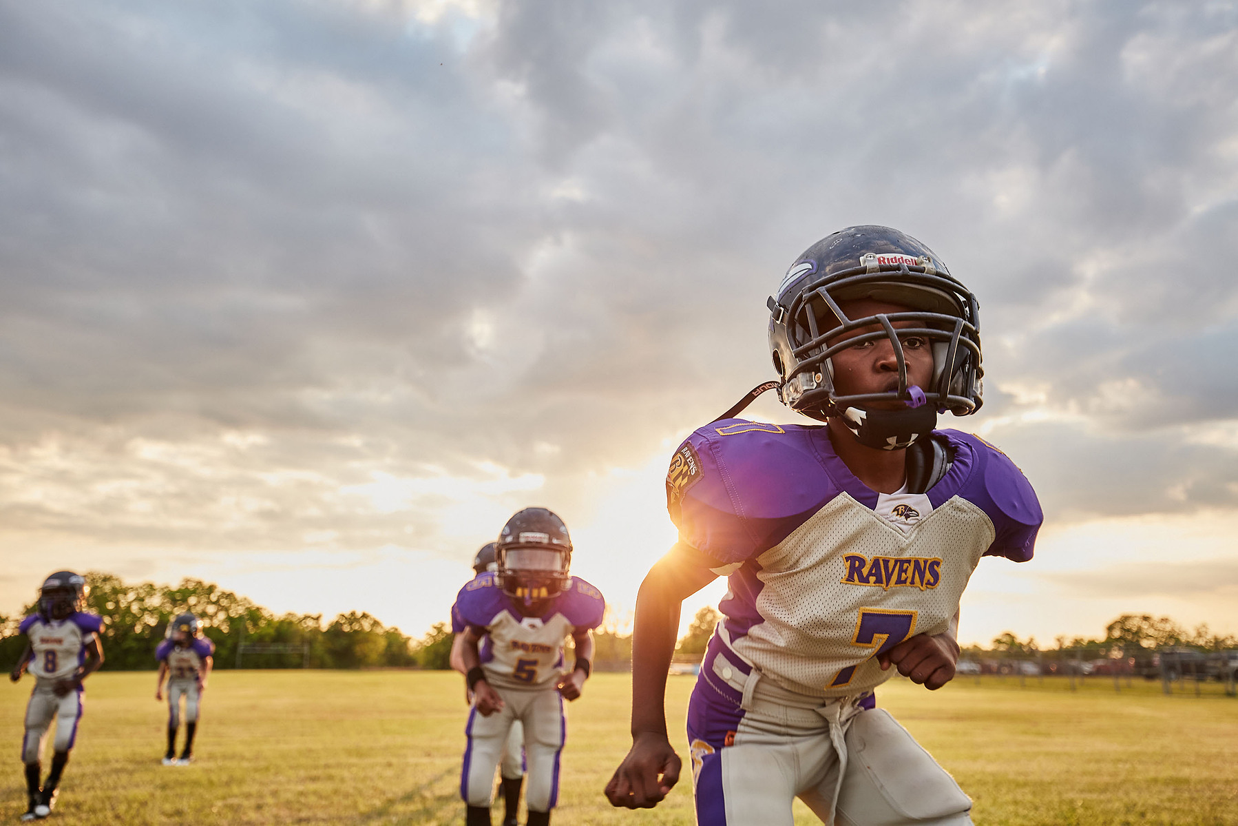 Young football players in white-and-purple uniforms run across a field in evening light.