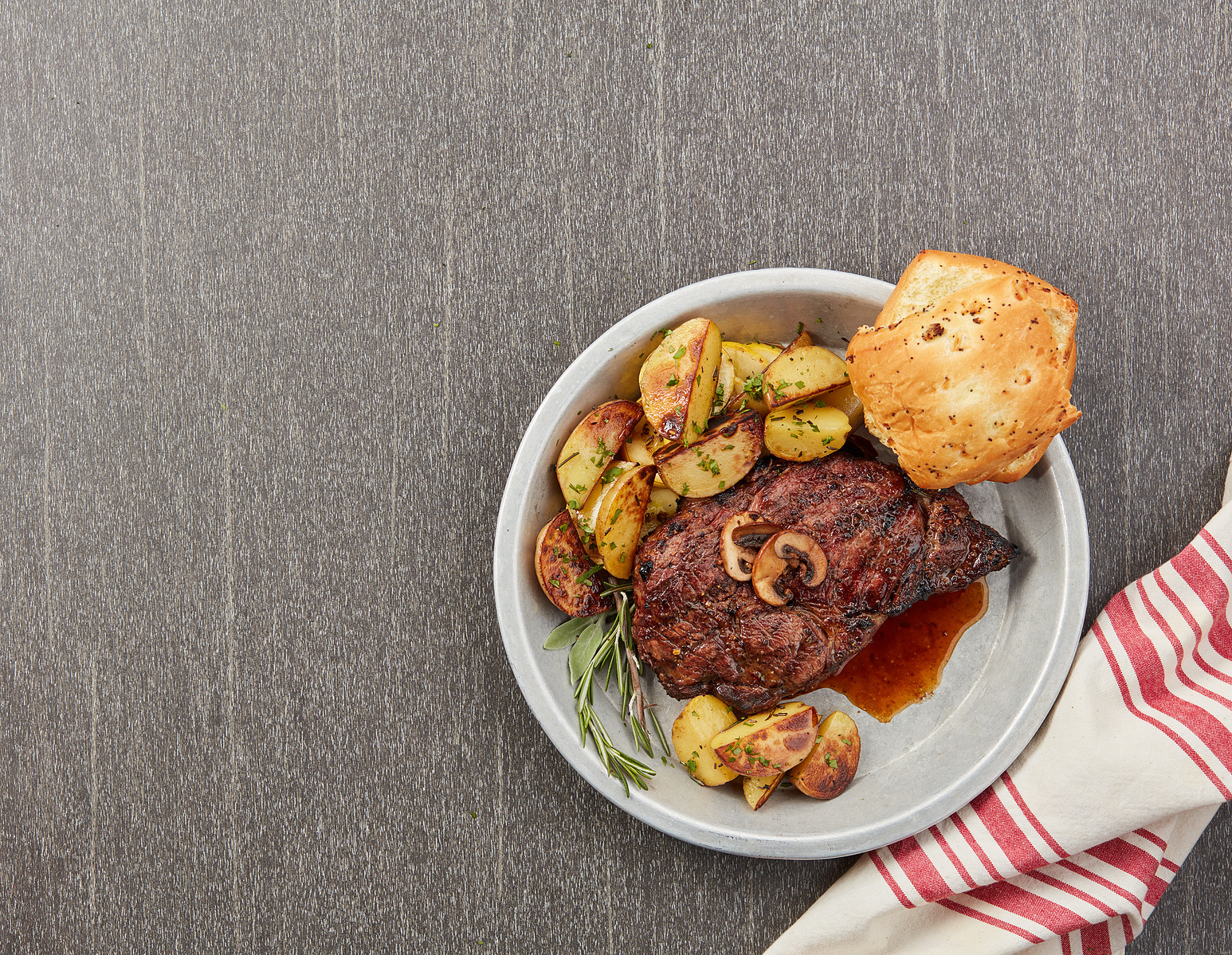 Glazed meat, roasted potato slices, and a bread roll on a white plate over a gray table.