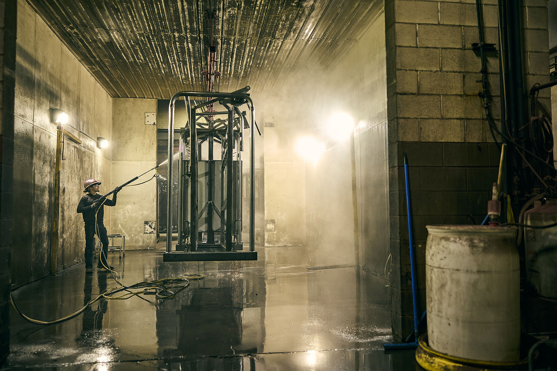 A worker sprays a suspended metal frame in a wet industrial wash bay.
