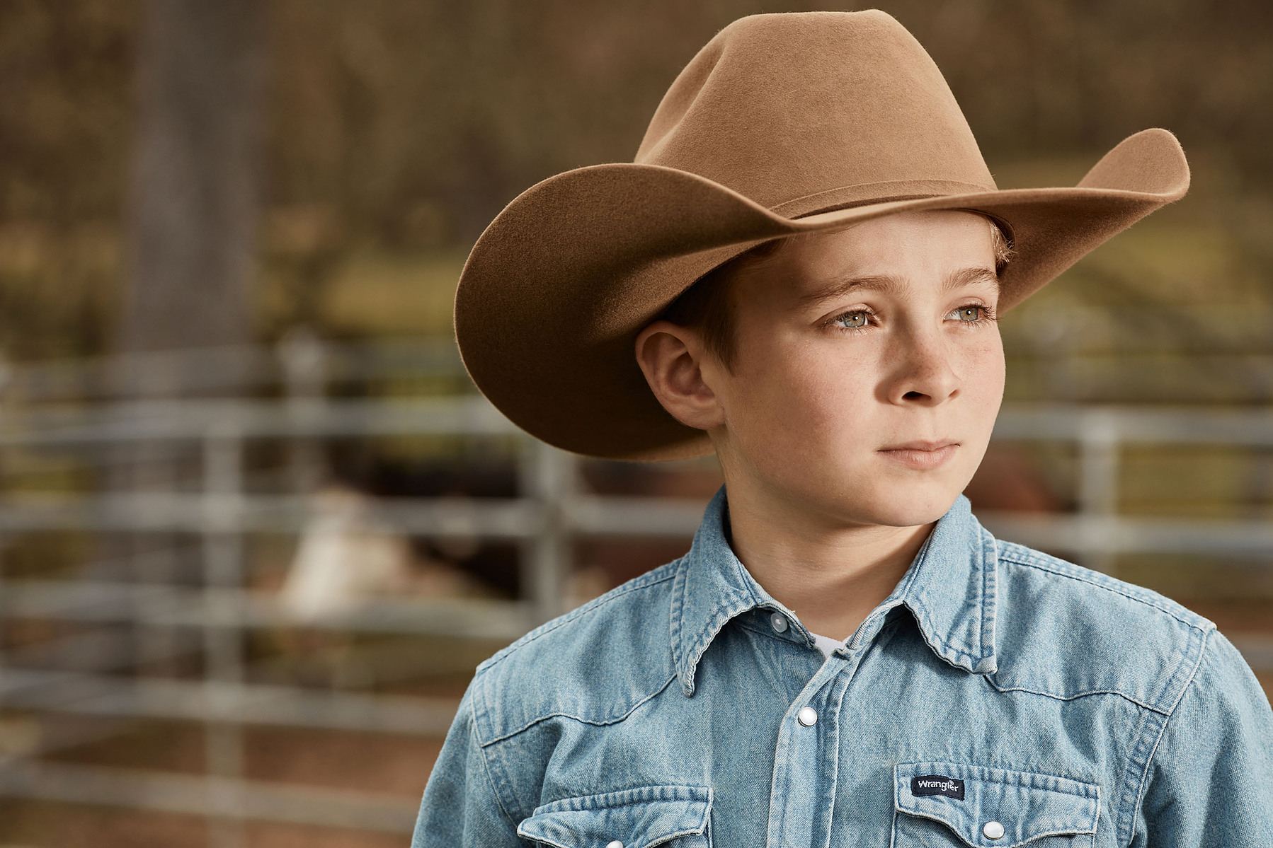 A child in a tan cowboy hat and denim shirt looks away from the camera outdoors.
