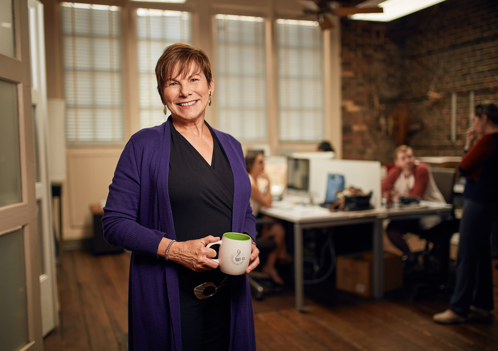 A smiling woman holding a mug stands in a brick-walled office with coworkers behind her.