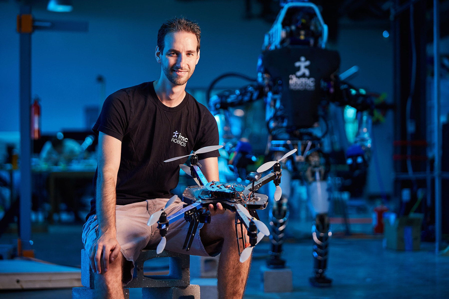 A smiling man holds a drone in a blue-lit workshop with larger equipment behind him.