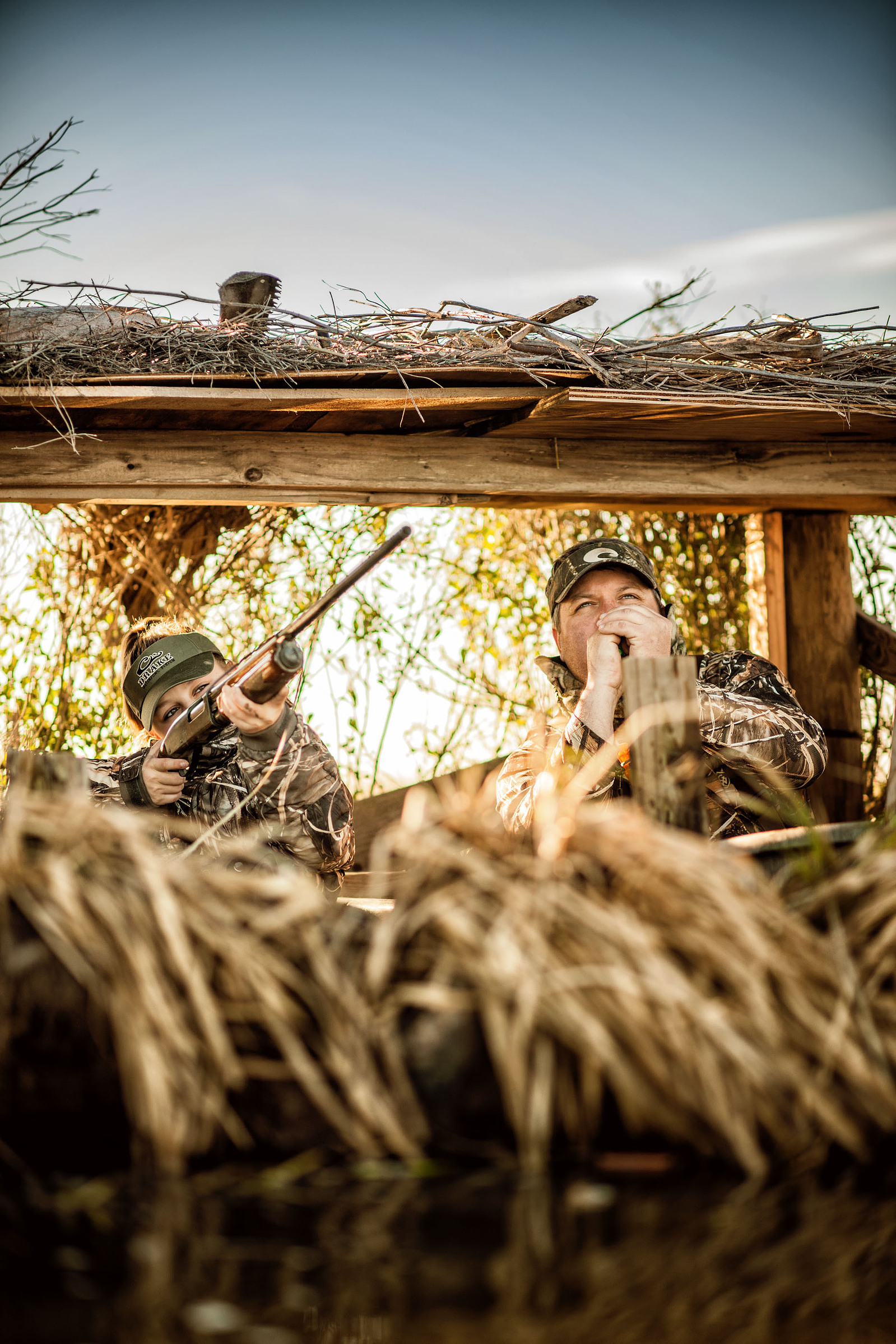 Two people in camouflage sit in a brush-covered hunting blind with a dog in the foreground.