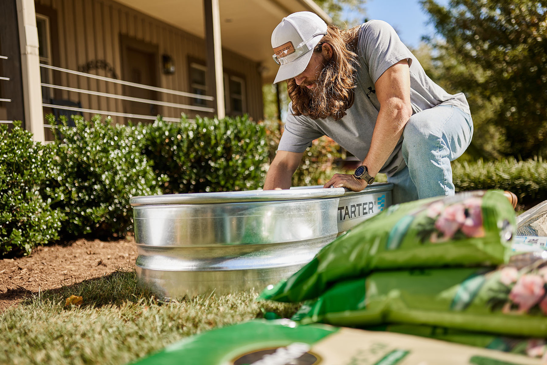 A person kneels beside a galvanized stock tank while arranging plants in a yard.