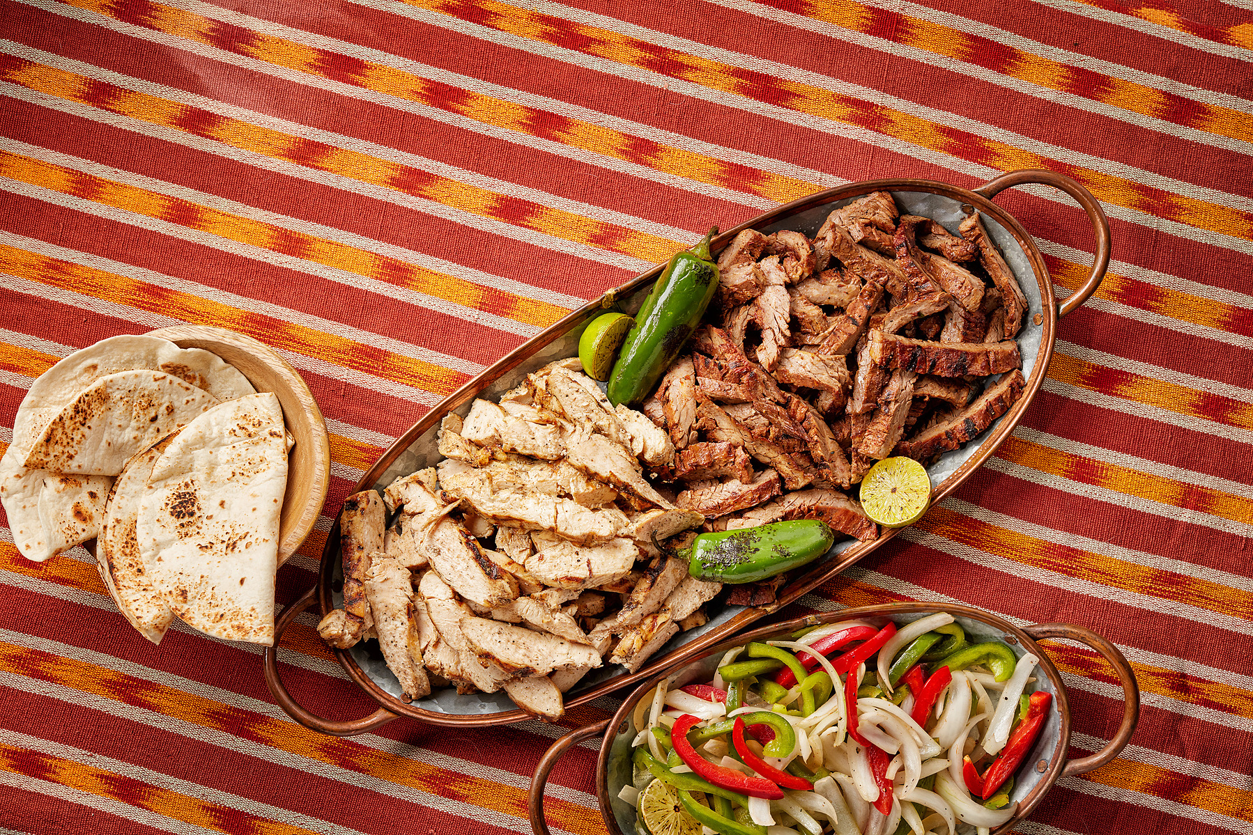 Grilled meat, peppers, onions, lime, and tortillas fill serving platters on a striped tablecloth.