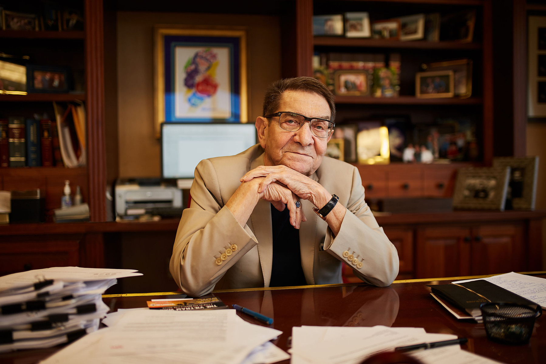 A man in glasses and a tan jacket sits at a desk surrounded by papers, books, and framed artwork.
