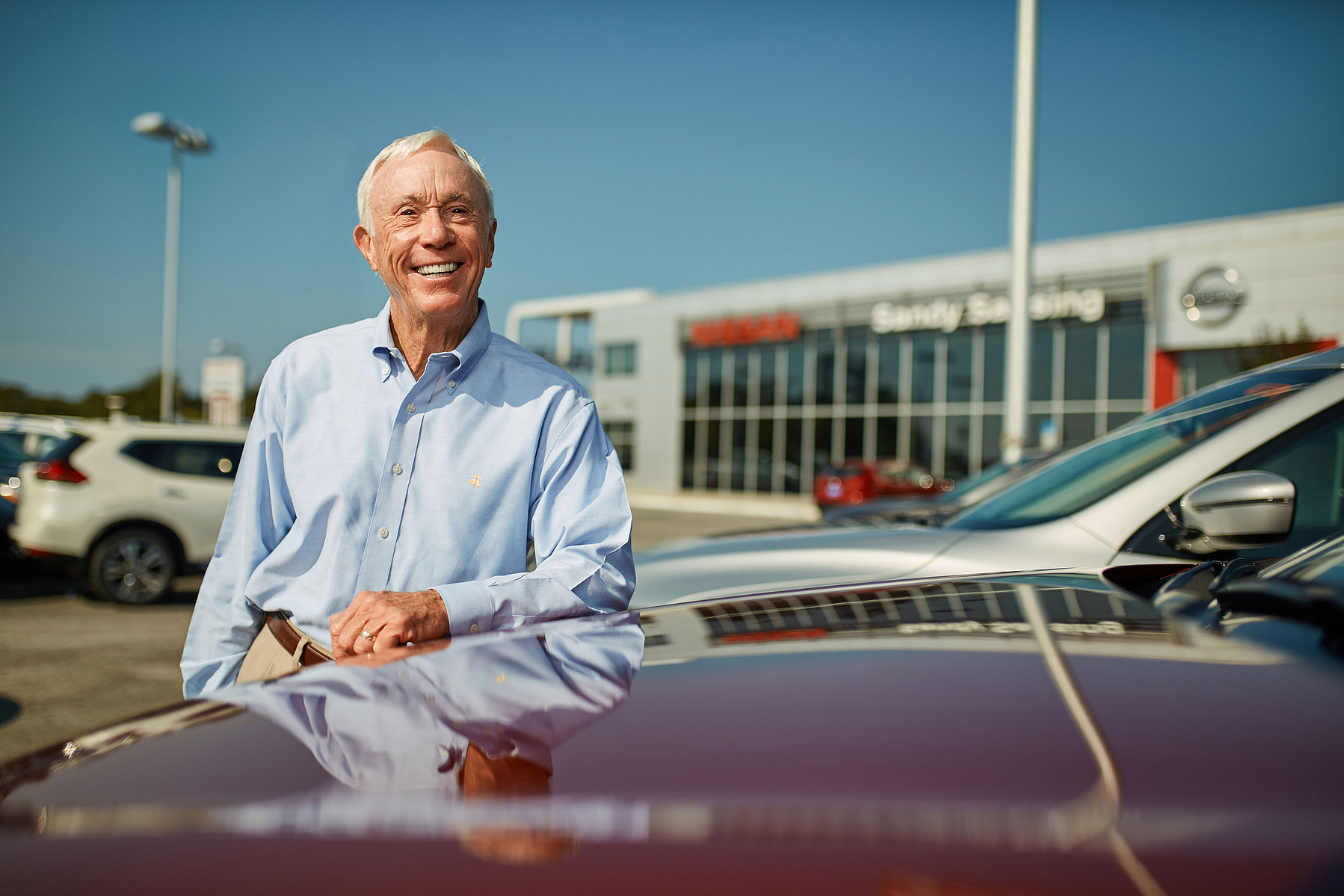 A smiling man leans beside a car outside an automotive dealership under a blue sky.