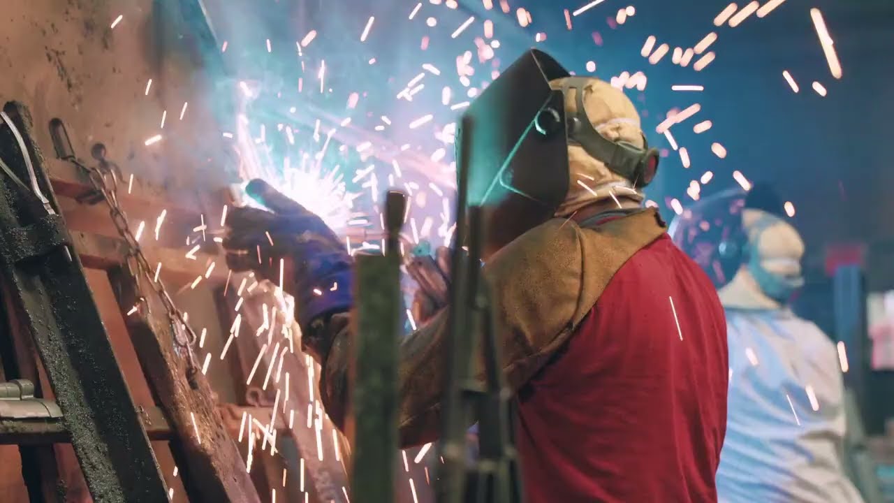 Film preview of a welder in protective clothing surrounded by sparks on a factory floor.