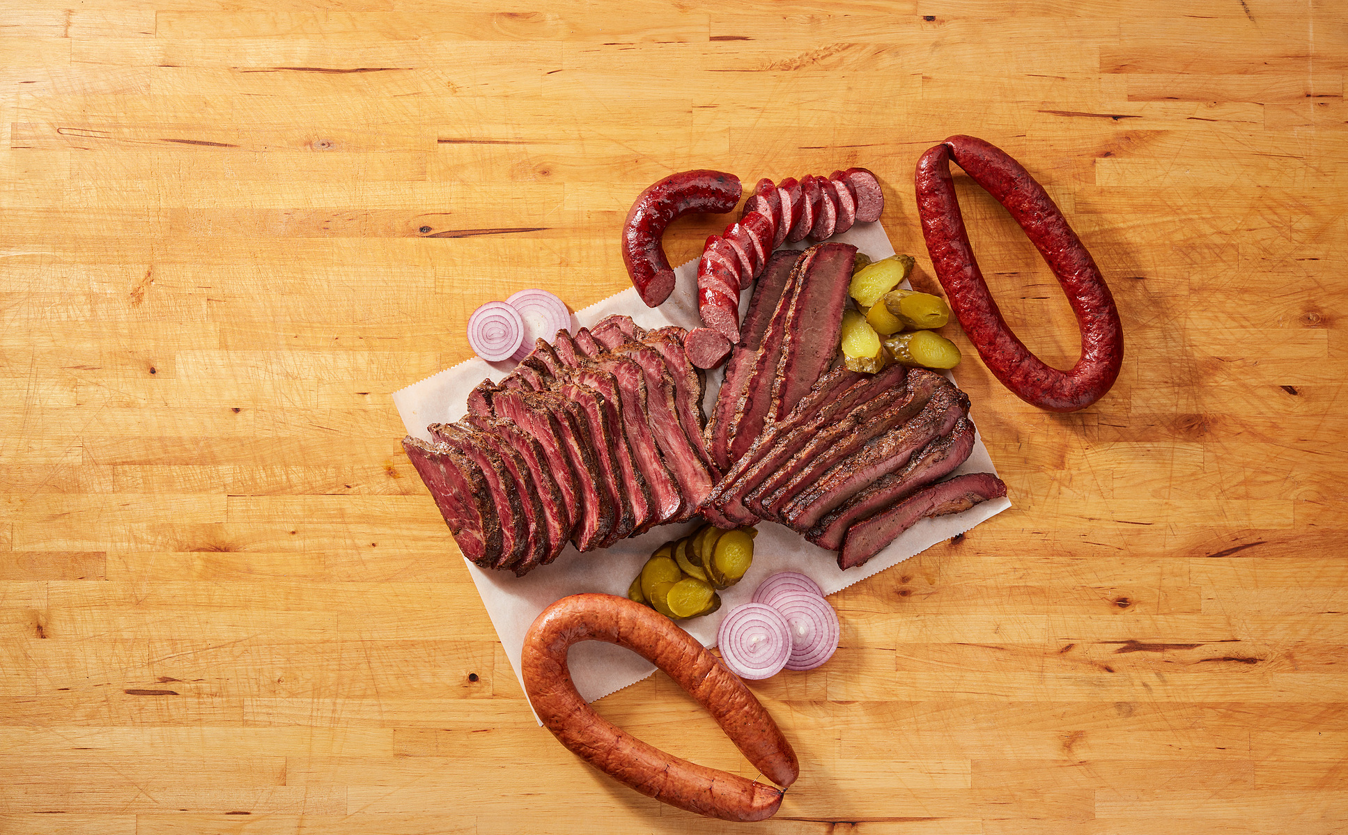 Sliced smoked meat, sausage, pickles, and red onion arranged overhead on a wooden table.