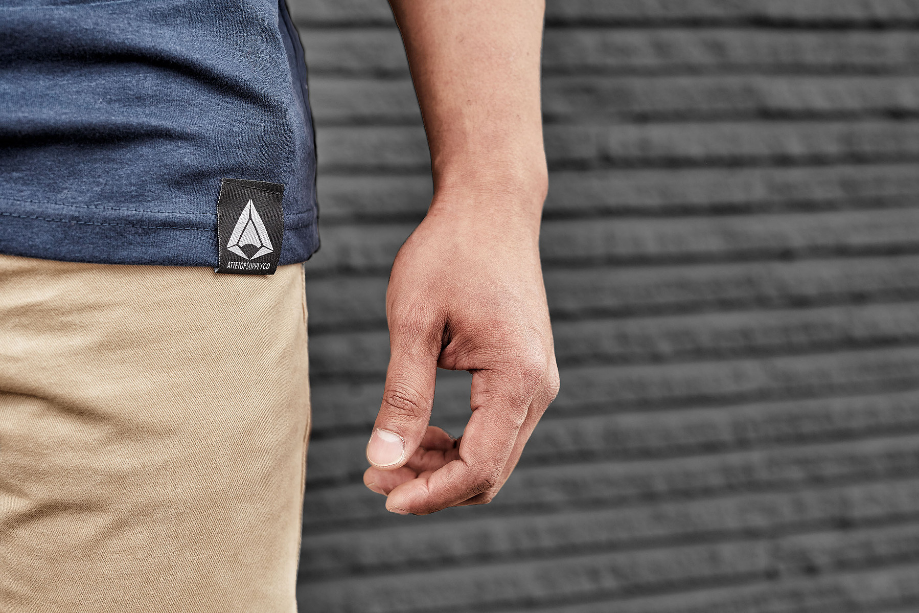 Close-up of a navy T-shirt hem, a hand, and tan trousers against a gray wall.