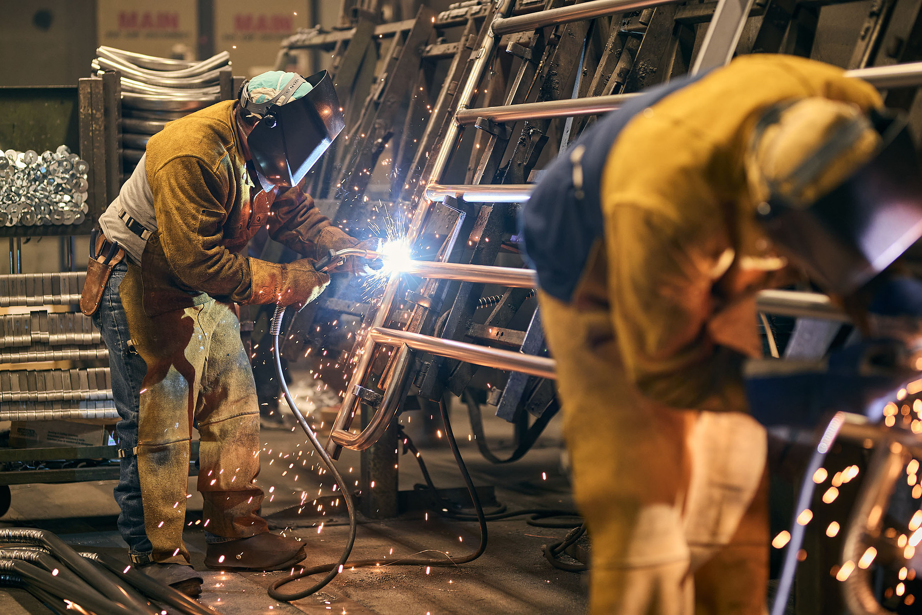 Two welders in protective helmets work on upright metal frames as sparks fly.