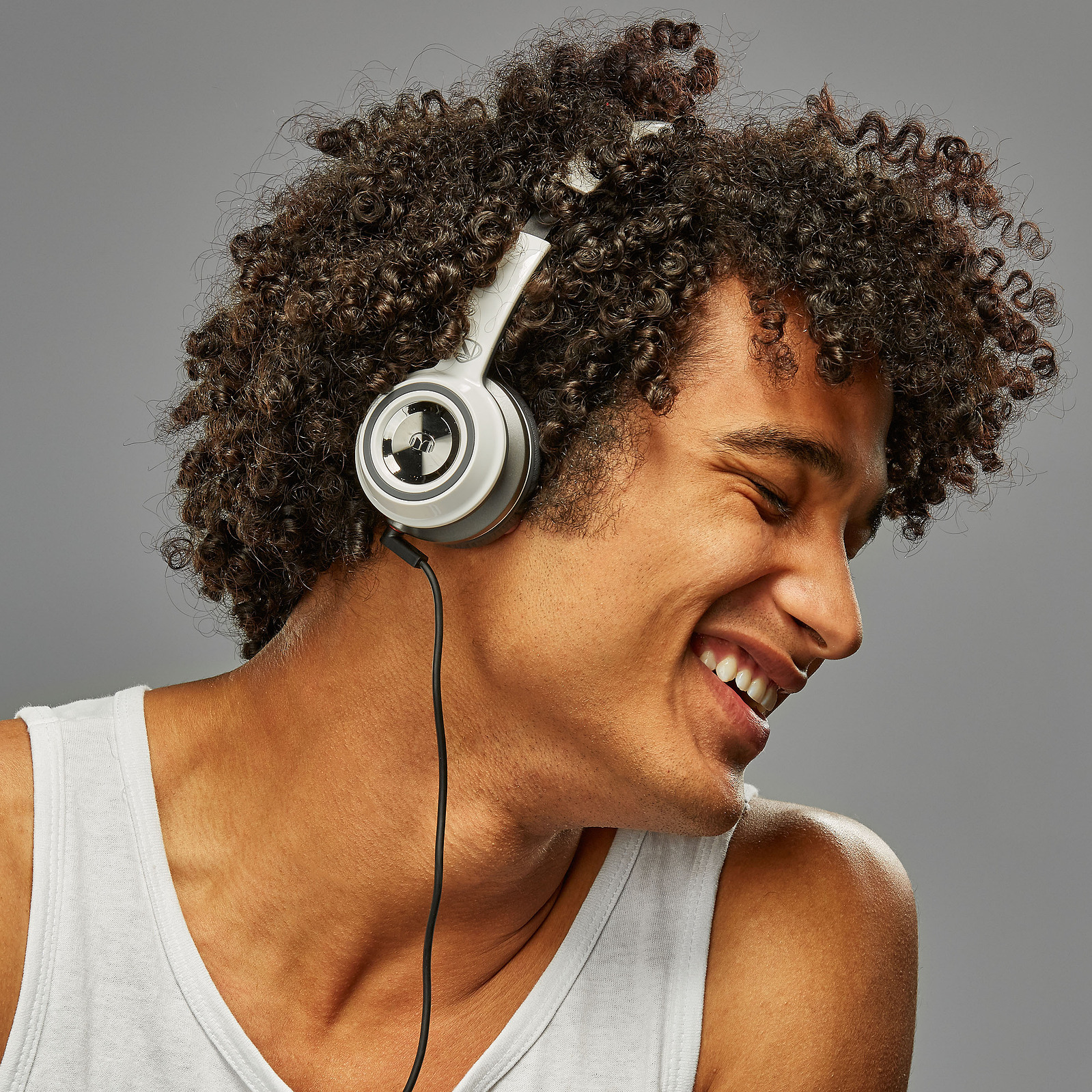 A smiling person with curly hair wears over-ear headphones against a gray background.