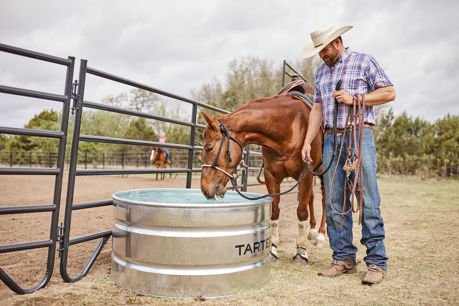 A cowboy holds a horse beside a galvanized water tank at a metal fence.