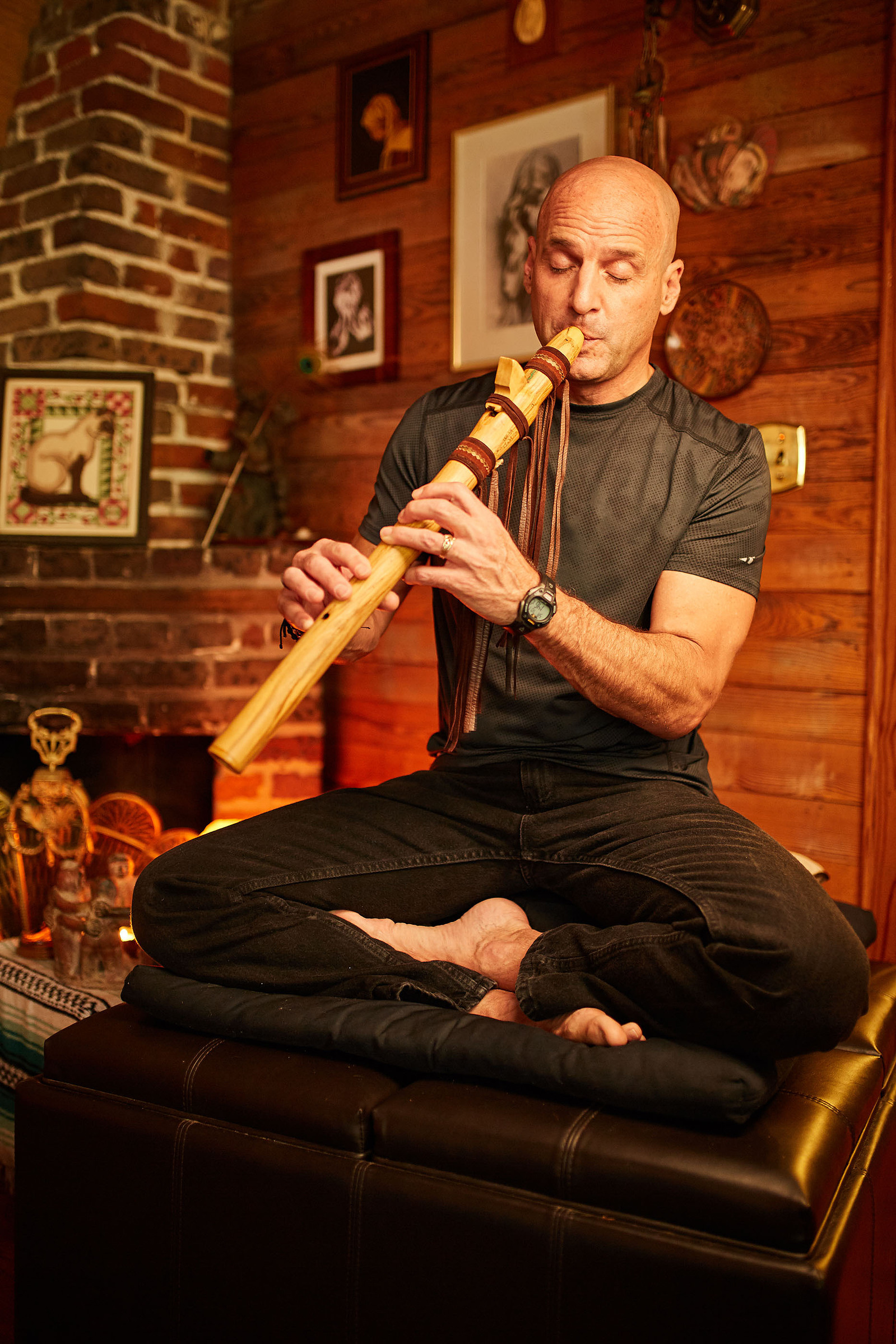 A seated man plays a wooden flute in a warmly lit room with brick walls and framed pictures.