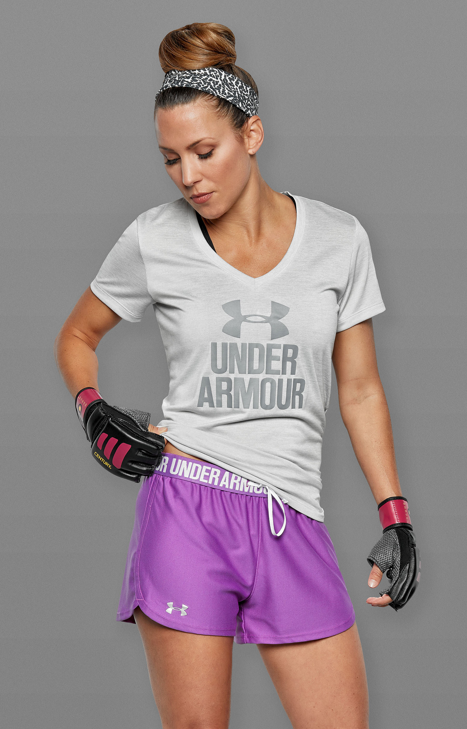 A woman in a gray Under Armour shirt, purple shorts, and training gloves adjusts her waistband.