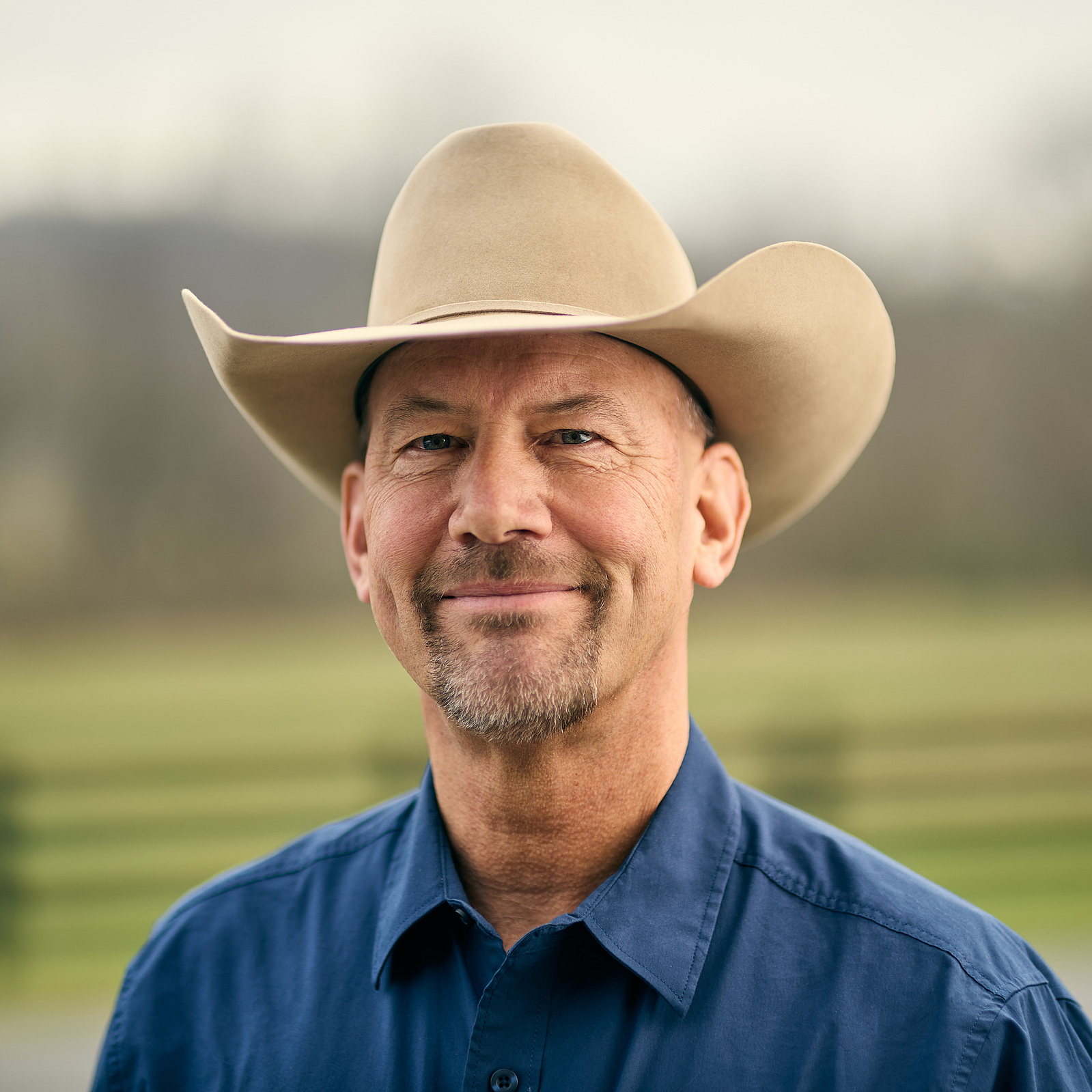 A smiling man in a pale cowboy hat and blue denim shirt photographed outdoors.