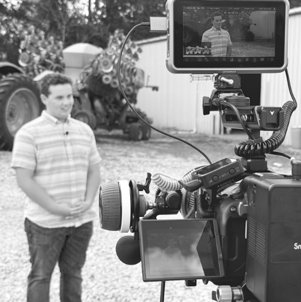 Behind-the-scenes view of a video camera filming a man outdoors near agricultural equipment
