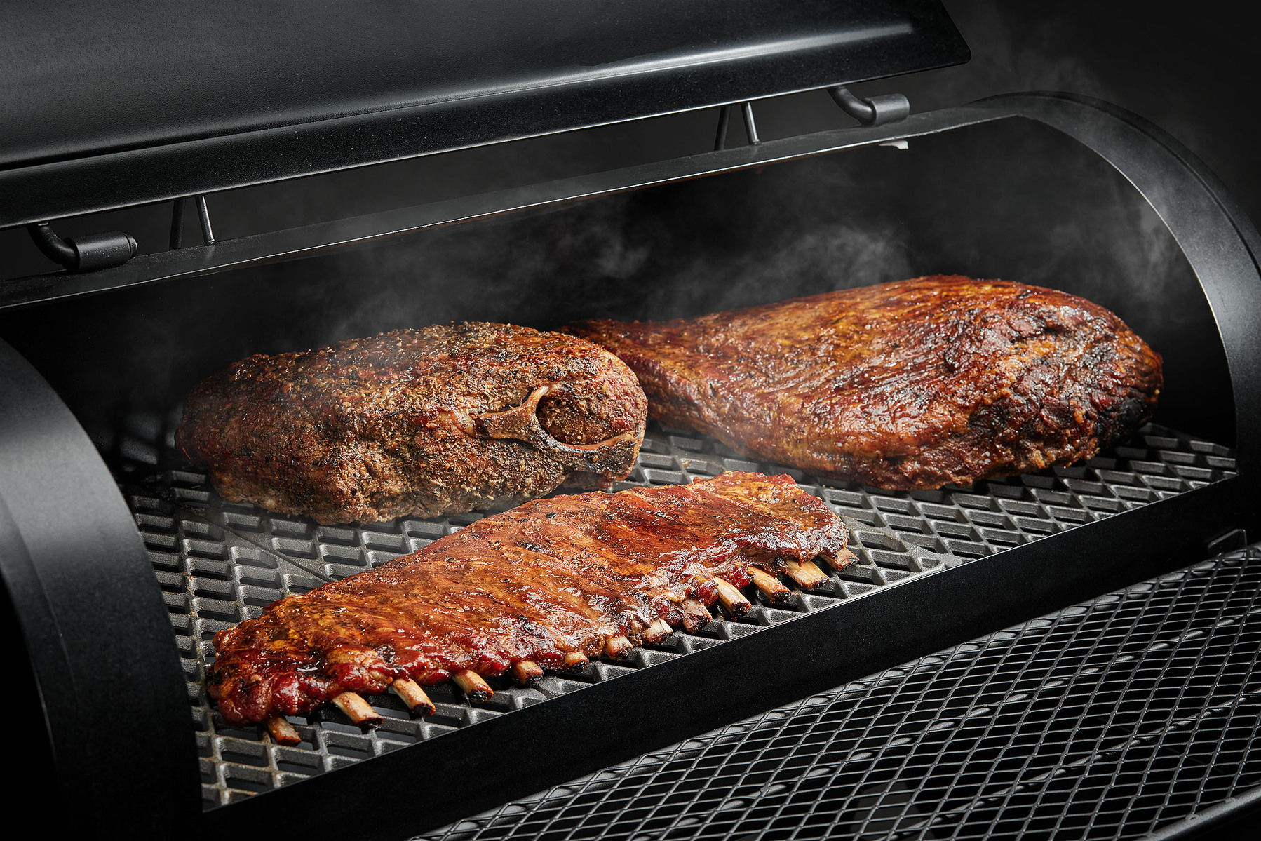 Large cuts of meat and a rack of ribs cook inside a dark barbecue smoker.