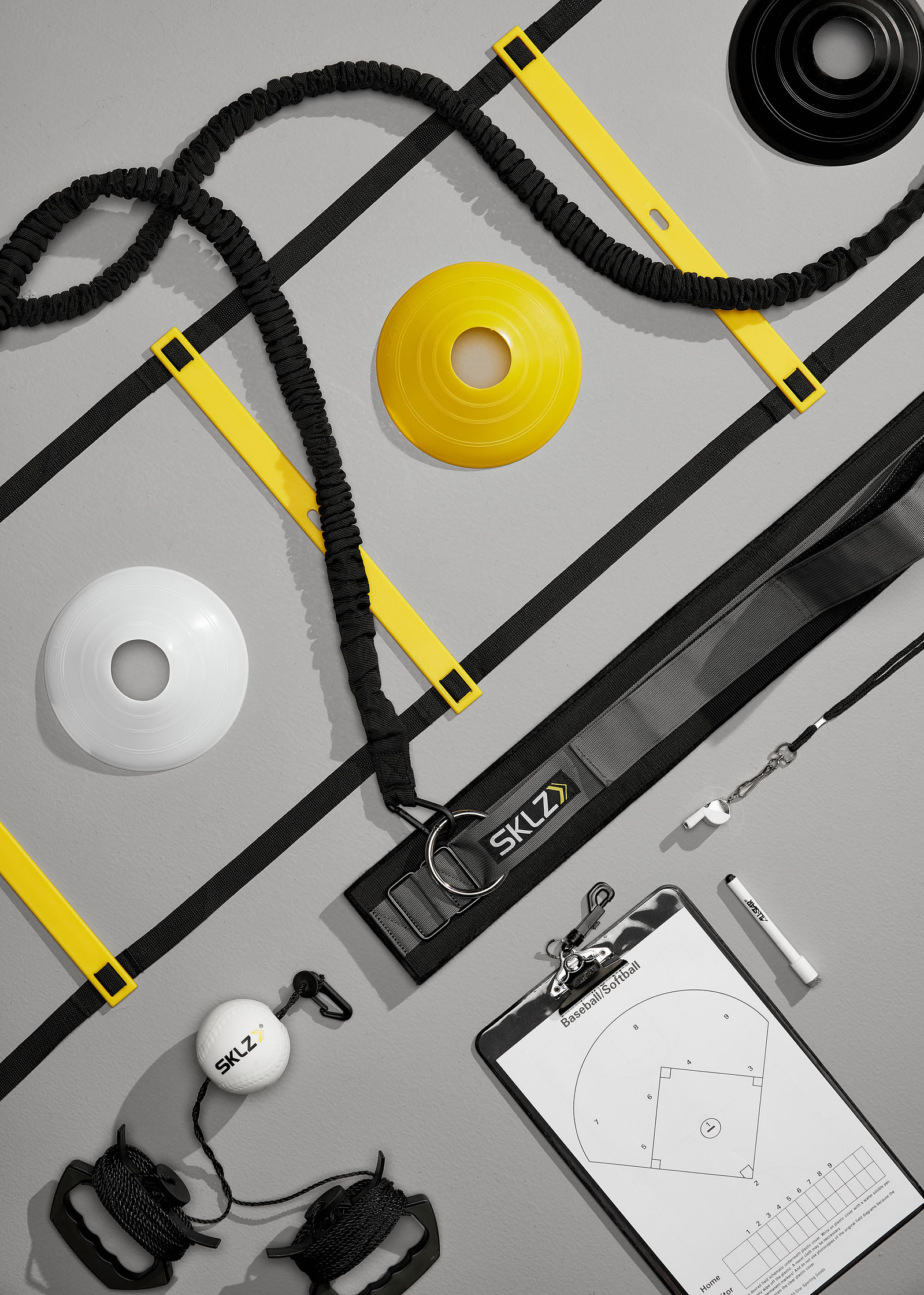An overhead arrangement of a yellow agility ladder, cones, resistance cords, a training belt, and a baseball practice clipboard on gray.