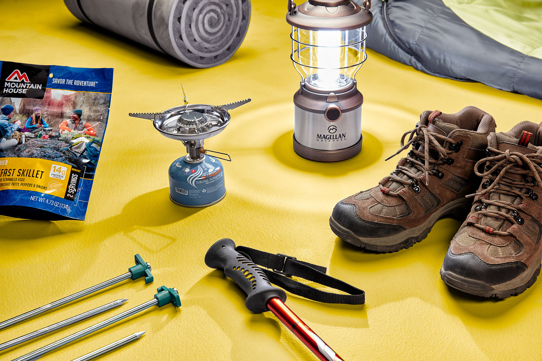 Camping equipment, hiking boots, poles, a stove, and a lantern arranged on a yellow surface.