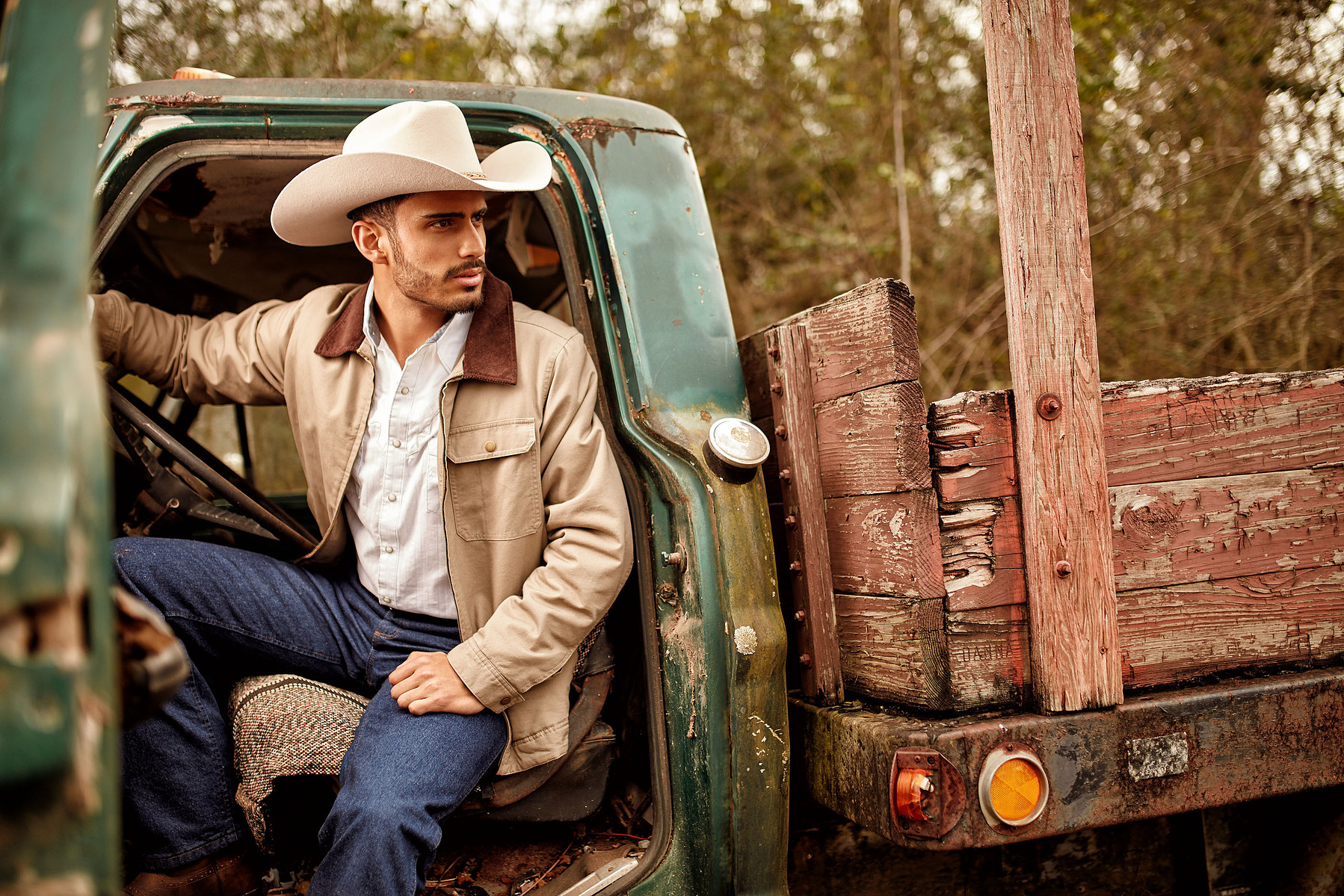 A man in a white cowboy hat and tan jacket sits in the open doorway of an older pickup truck.